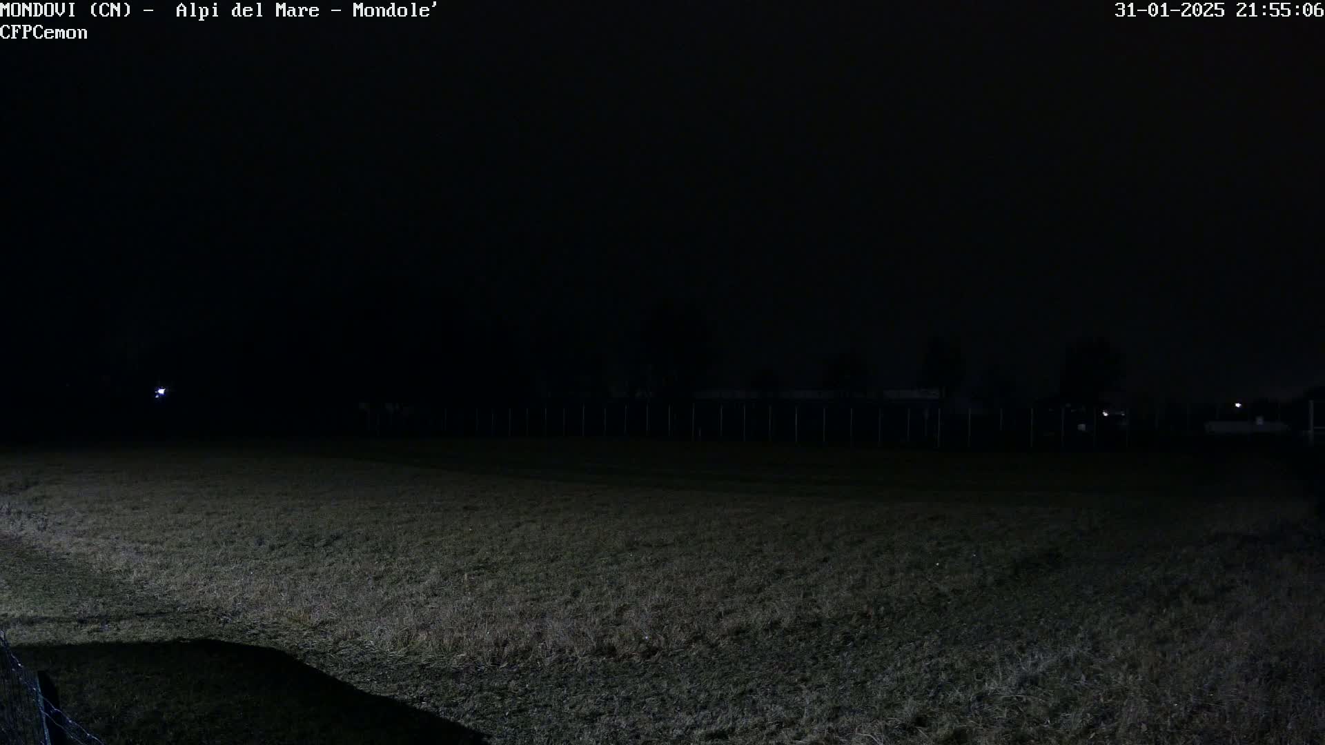 A dark, grassy field is visible at night under a mostly clear sky, with a fence and dimly lit structures in the background.