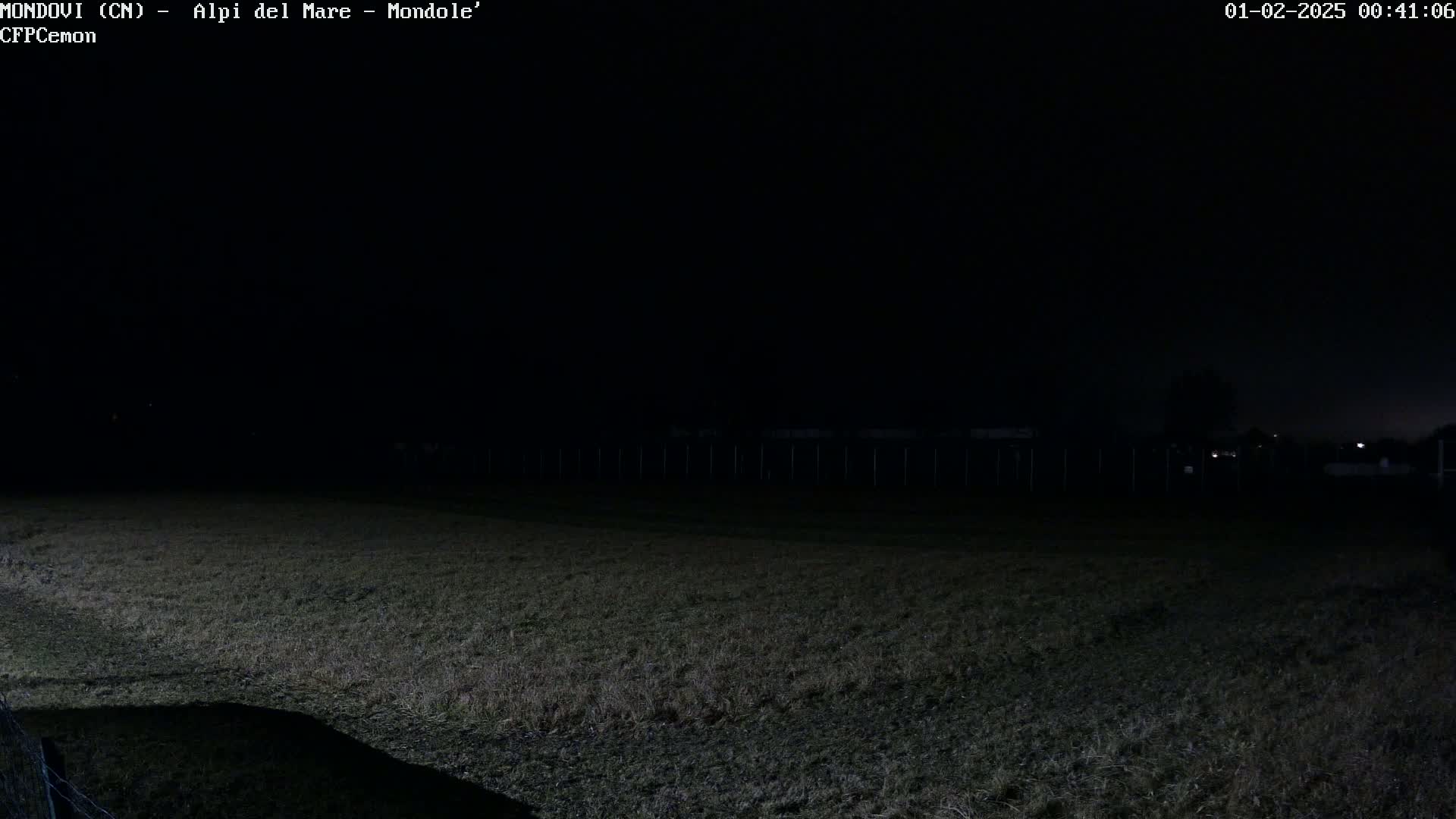 A dark, nighttime view of a grassy field, enclosed by a fence, with a few distant lights visible in the background.