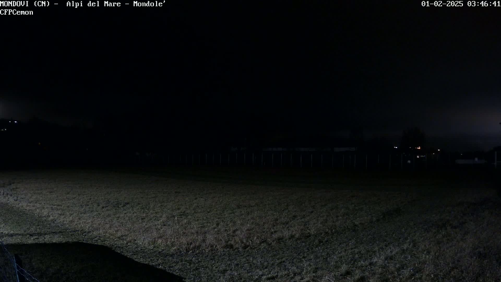 A dark, nighttime scene shows a grassy field, a fence, and dimly lit buildings in the background under a cloudy sky.