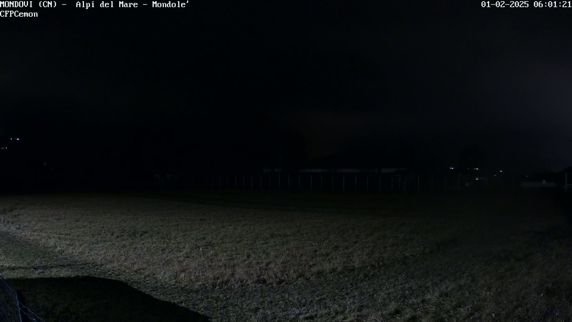 A dark, nighttime field is visible under a cloudy sky, with a fence and dimly lit structures in the distance.