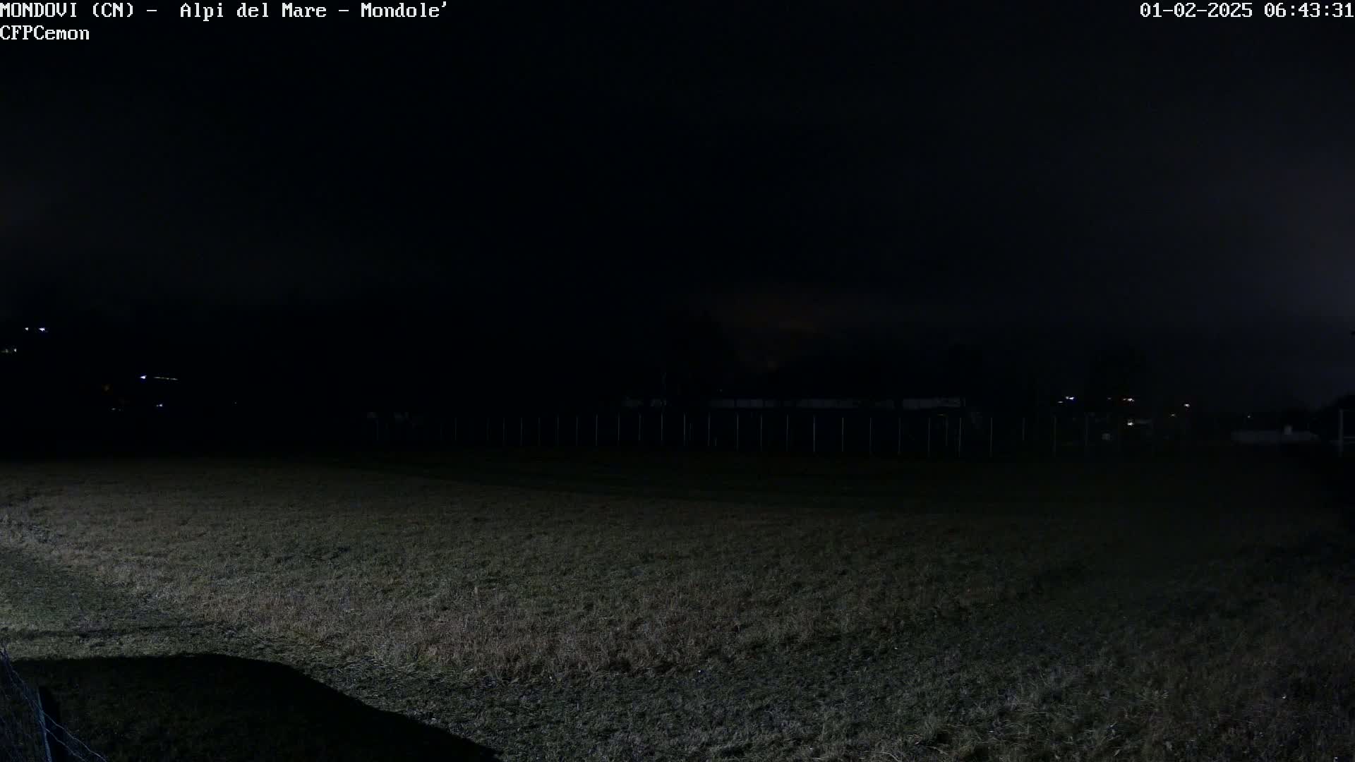 A dark, nighttime view of a grassy field, bordered by a fence, with a few distant lights visible under a mostly clear, dark sky.