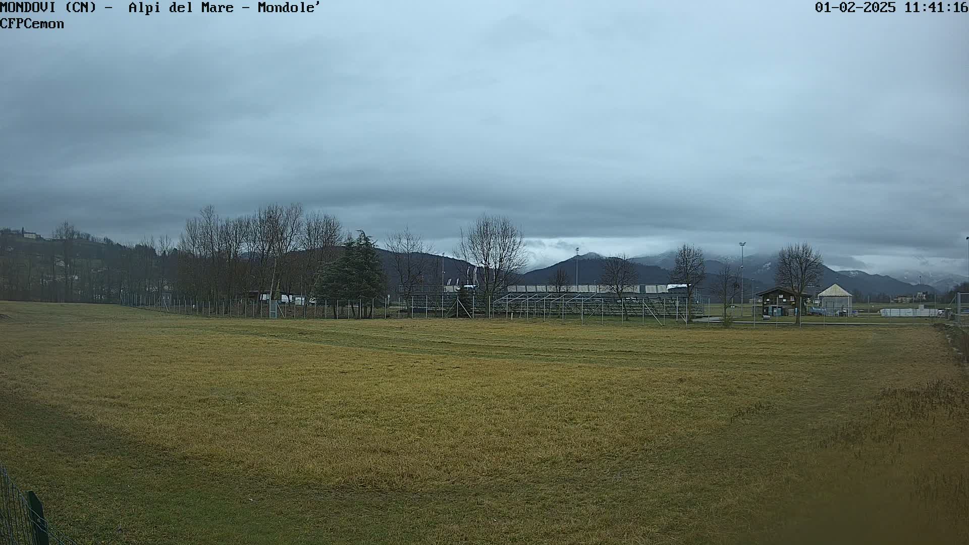 A large, empty field stretches towards bleachers and distant, snow-capped mountains under an overcast sky.