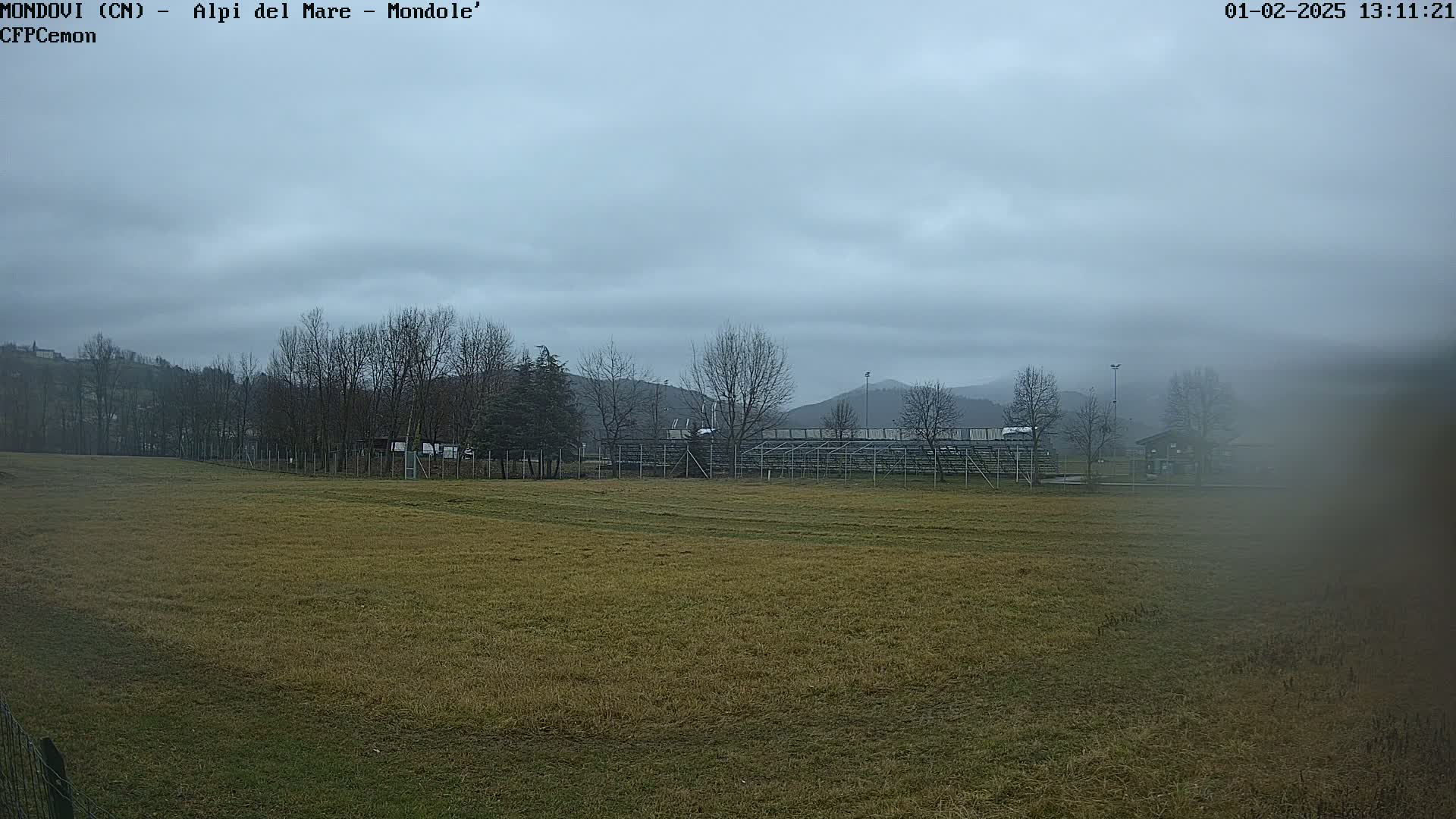 A large, mostly brown field stretches toward bleachers and distant hills under a cloudy sky.