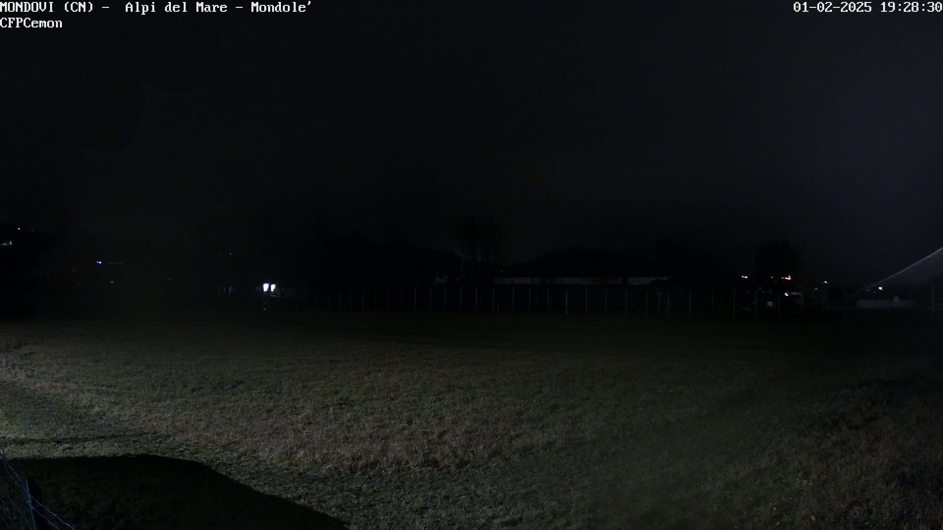 A dark, nighttime view of a grassy field, with a few distant lights visible beyond a fence.