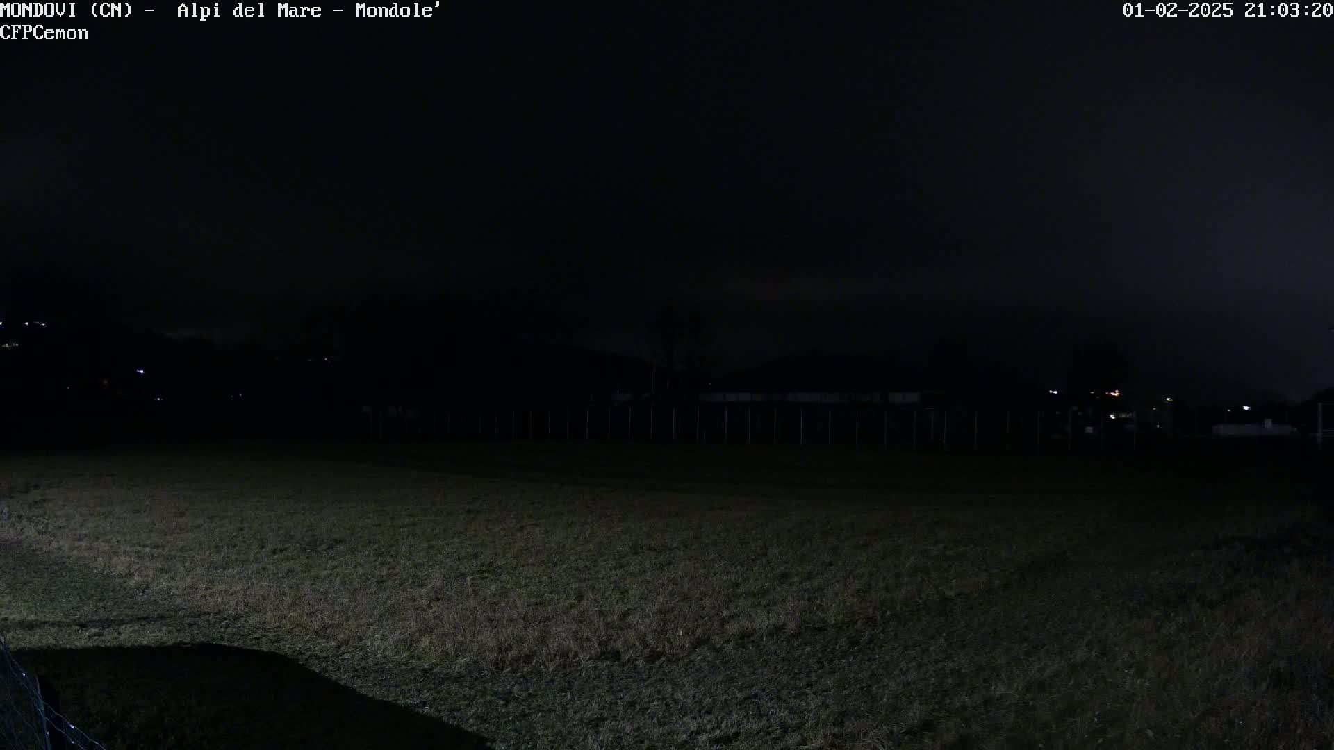 A dark, nighttime view of a grassy field extending to a fence and distant, dimly lit structures under a mostly cloudy sky.