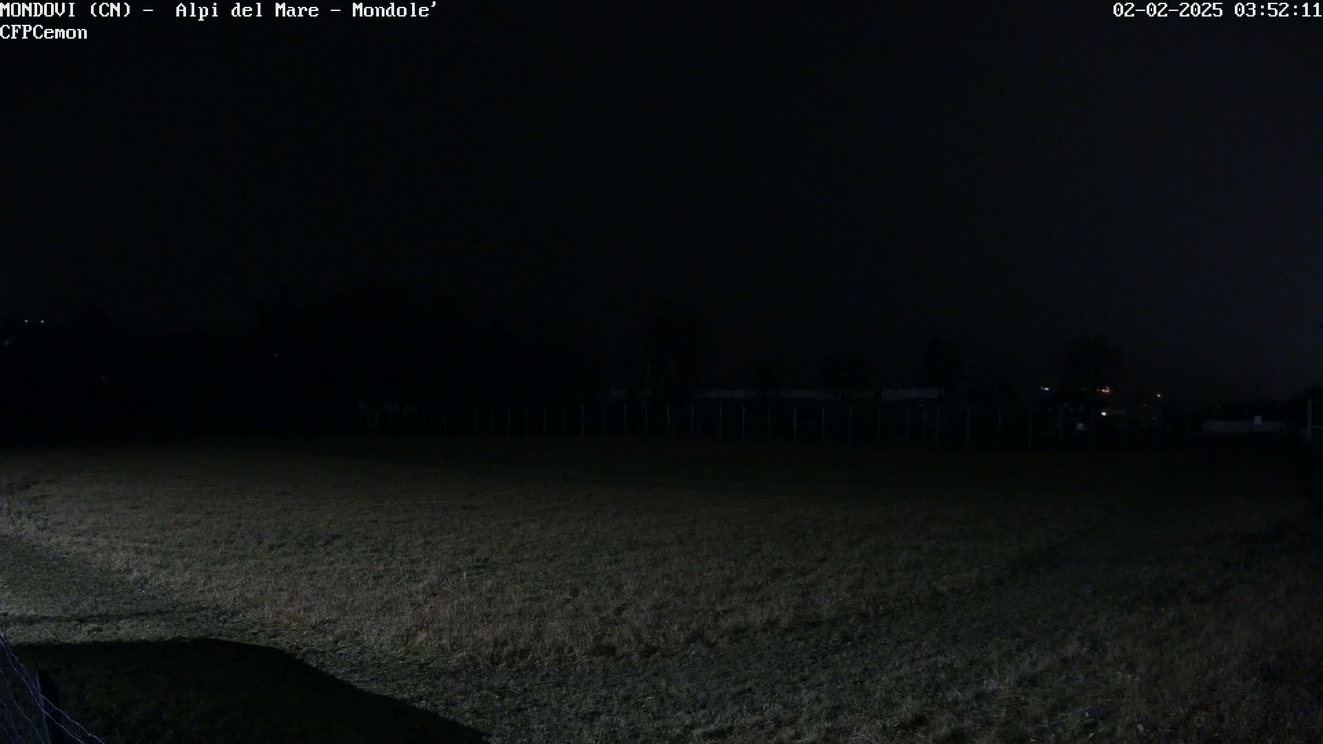 A dark, nighttime view of a grassy field enclosed by a fence, with dimly lit structures visible in the background under a mostly clear sky.