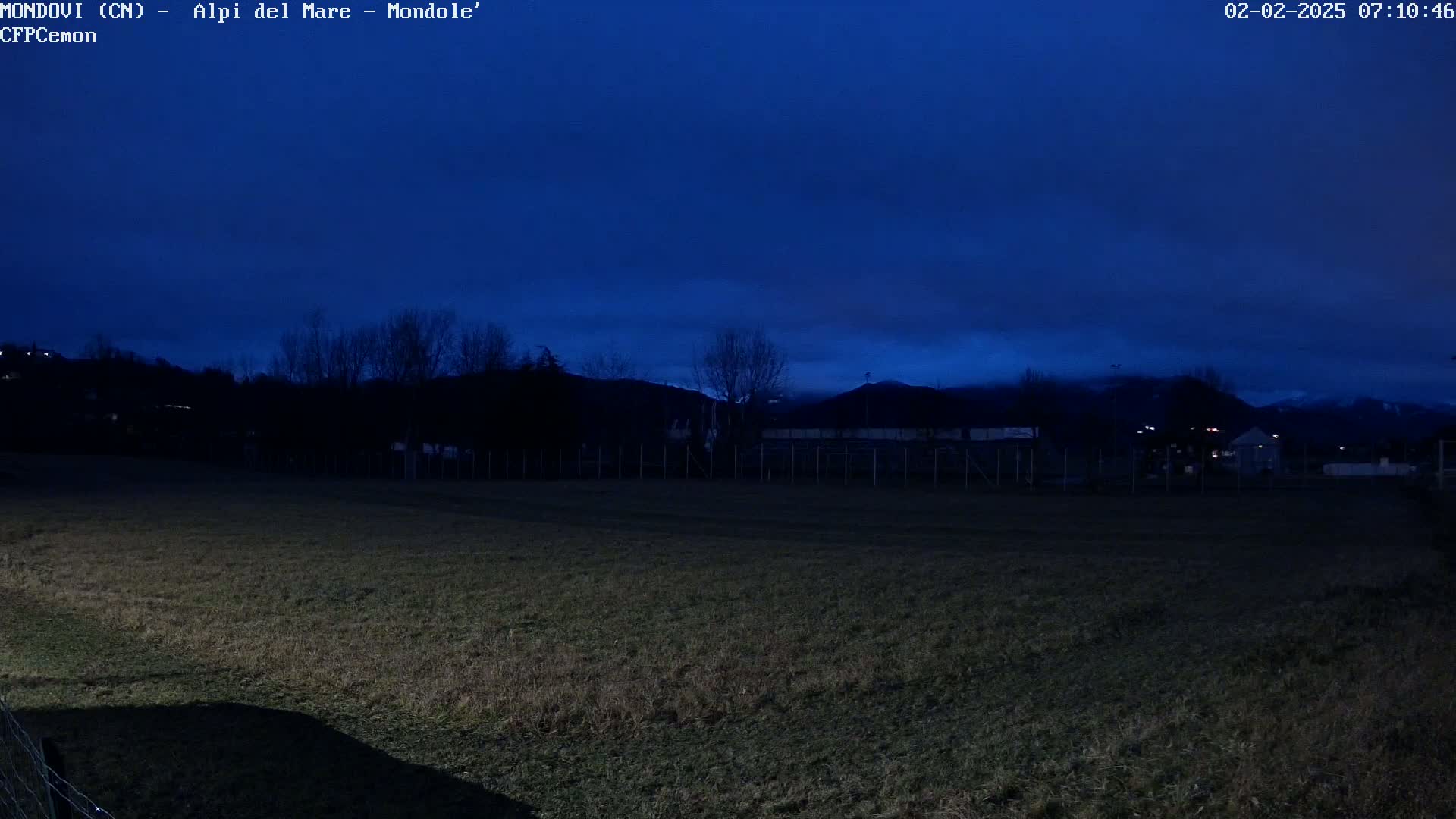 A dark, nighttime view of a grassy field, enclosed by a fence, with silhouetted trees and distant hills under a deep blue, mostly clear sky.