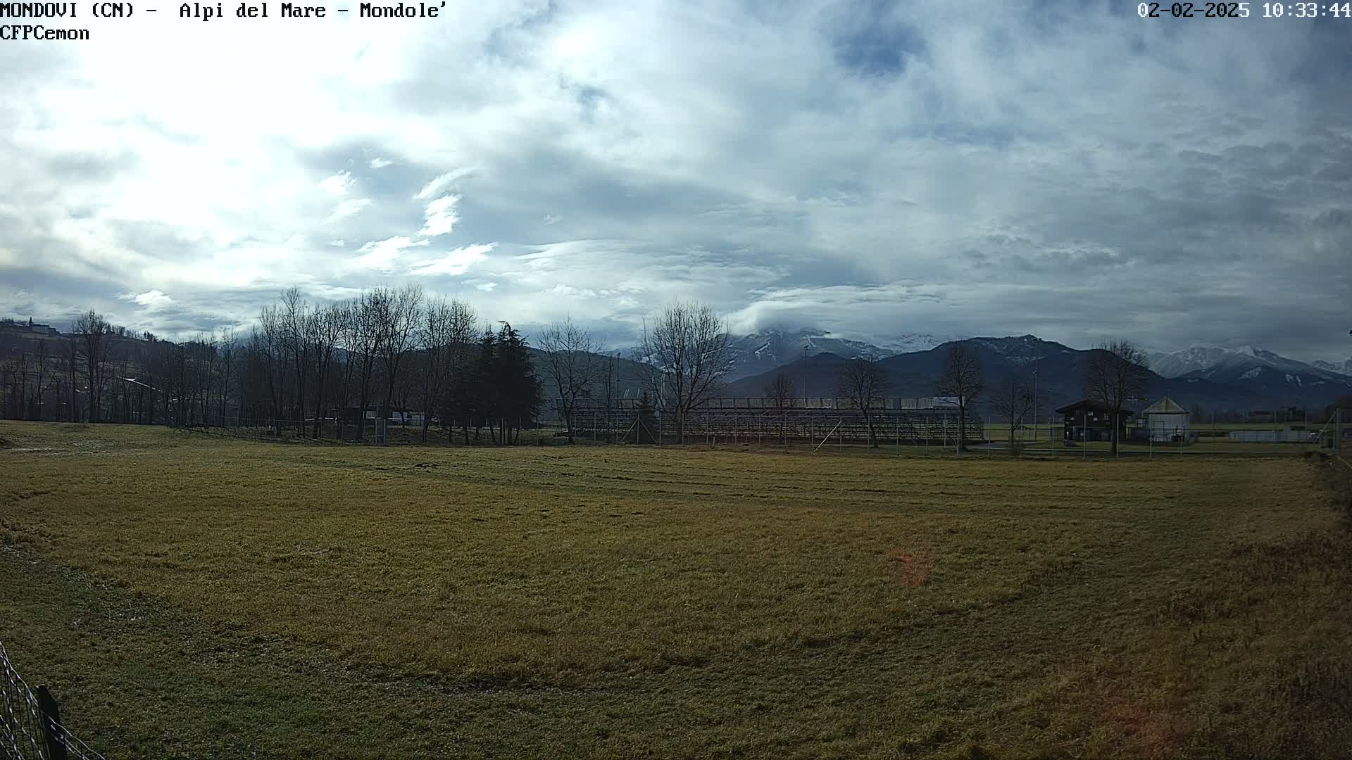 A mostly cloudy day reveals a snow-capped mountain range in the distance behind a field with leafless trees and an empty bleacher structure.
