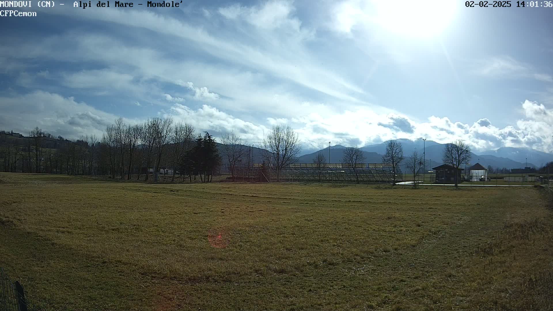 A partly cloudy day reveals a grassy field, bleachers, and a small building, all set against the backdrop of distant mountains.