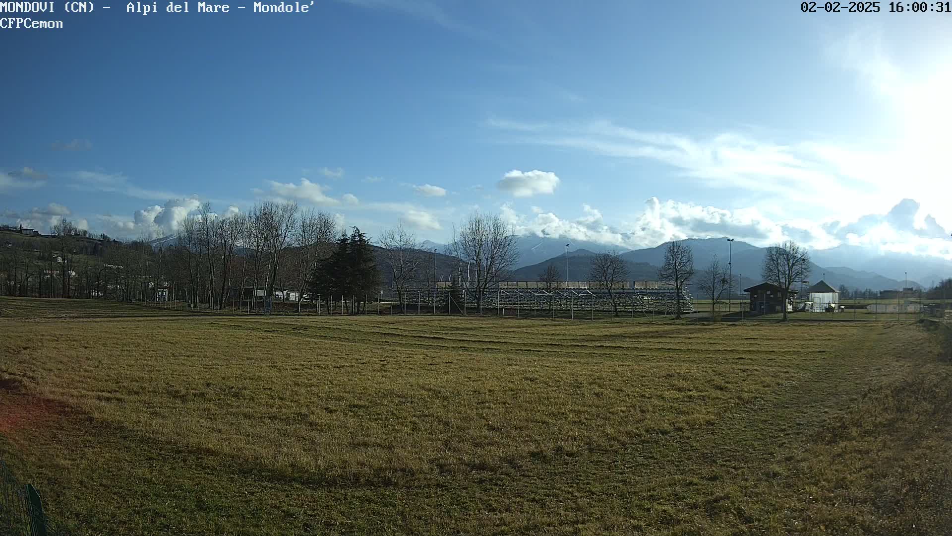 A mostly sunny day shows a grassy field with a few bare trees, a small building, and bleachers in the background, all set against a backdrop of mountains under a partly cloudy sky.