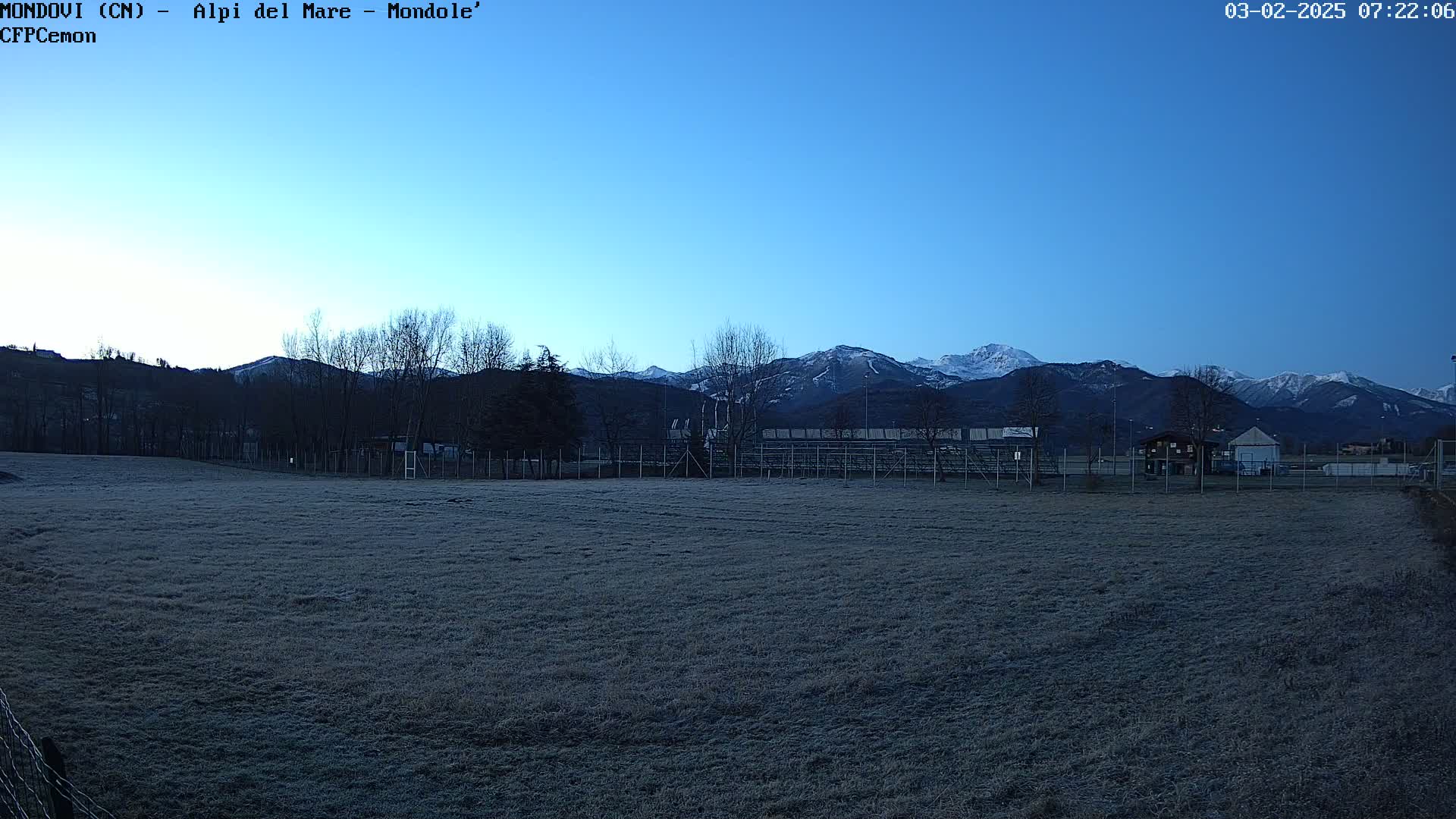 A frost-covered field sits before a fenced-in sports area, with snow-capped mountains visible in the background under a clear, twilight sky.
