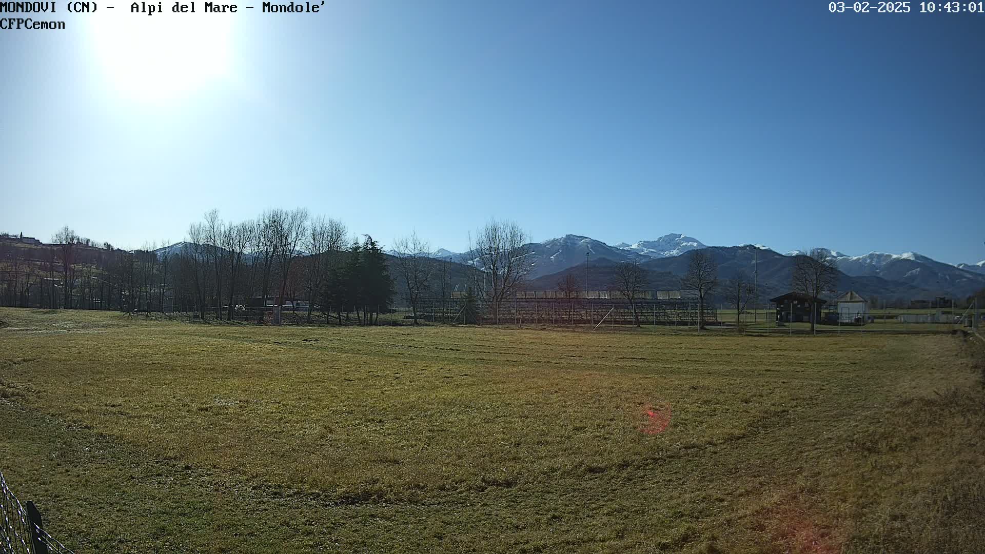 A brown field, sparsely covered with grass, stretches out before snow-capped mountains under a clear blue sky.