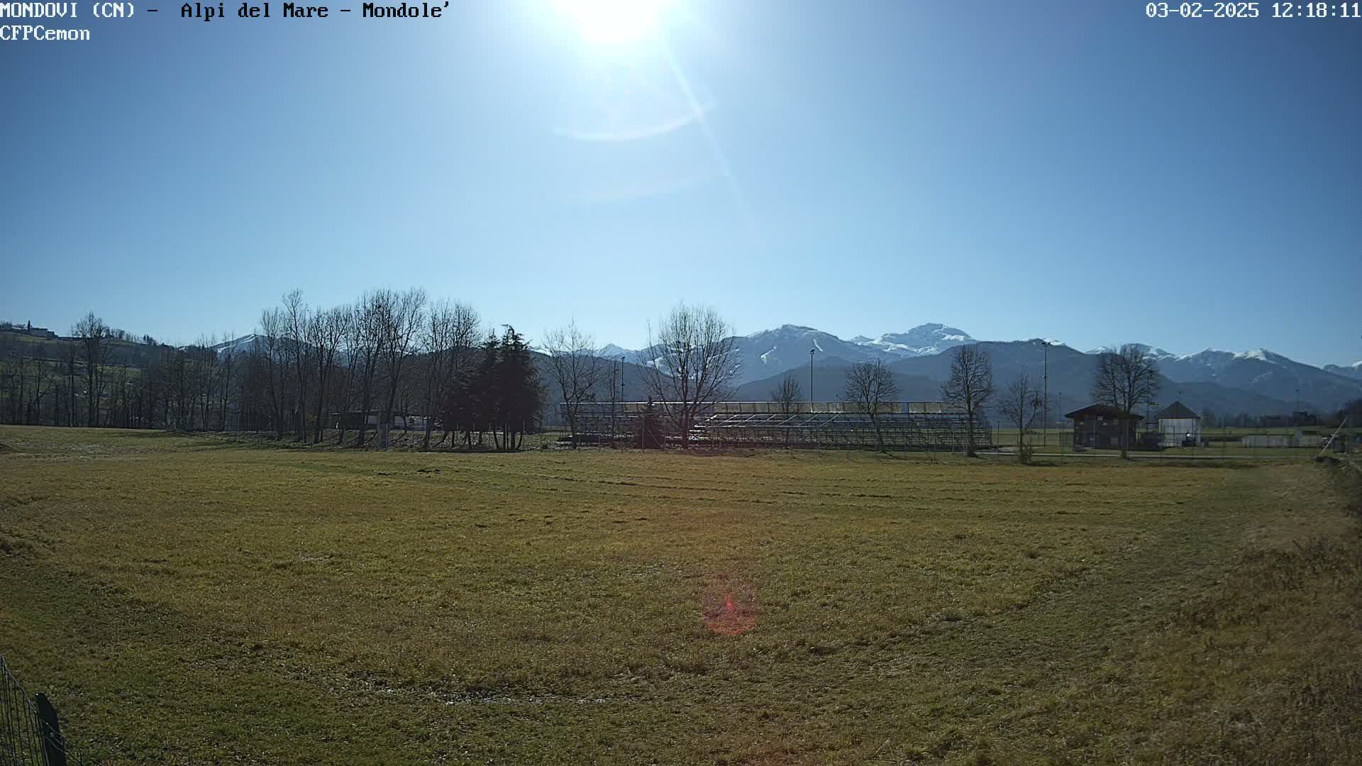 A sunny day reveals a grassy field with a metal bleacher structure, bare trees, and snow-capped mountains in the distance.
