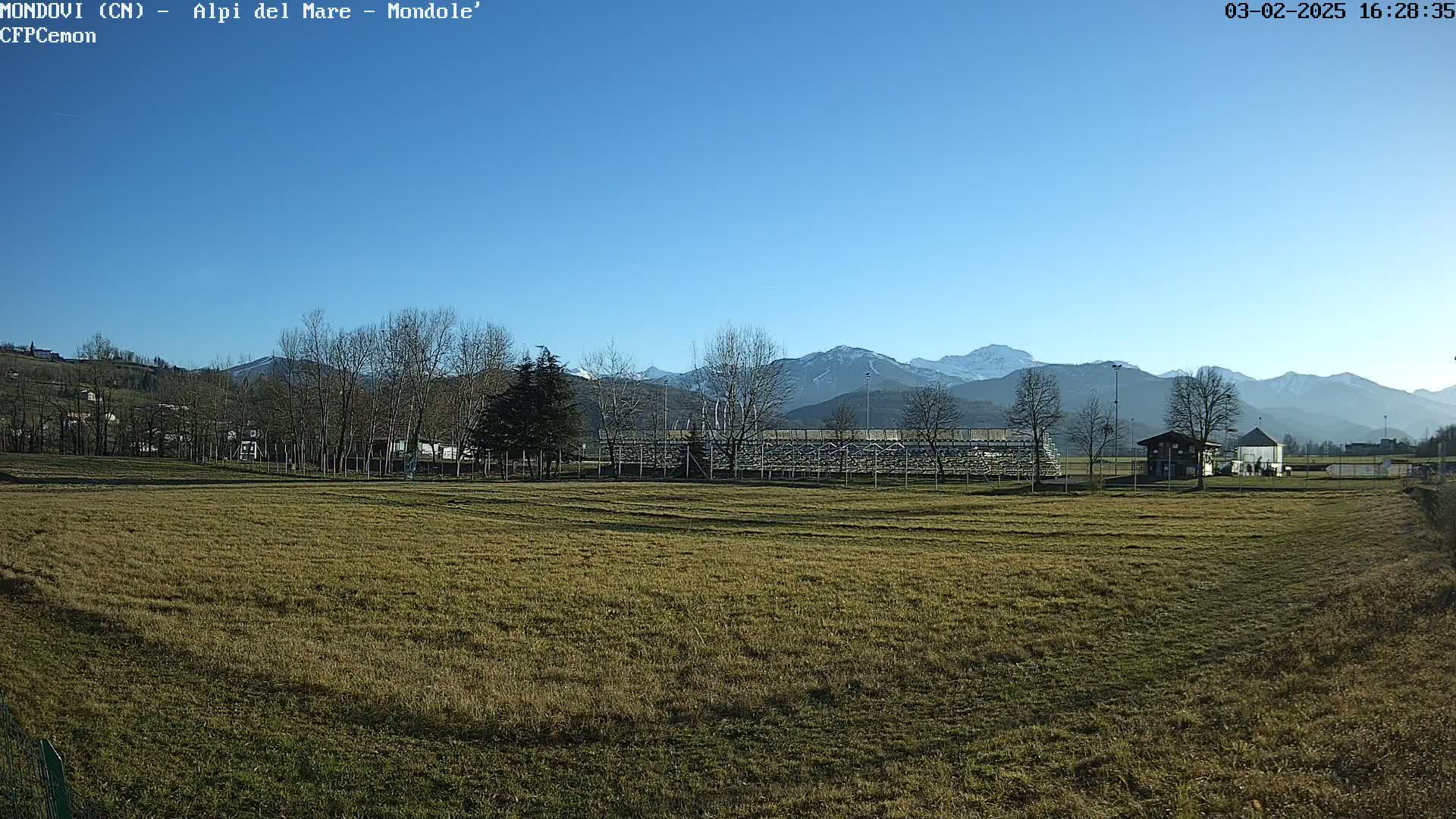 A grassy field with a small building and bleachers is situated in the foreground, while snow-capped mountains rise in the background under a clear blue sky.