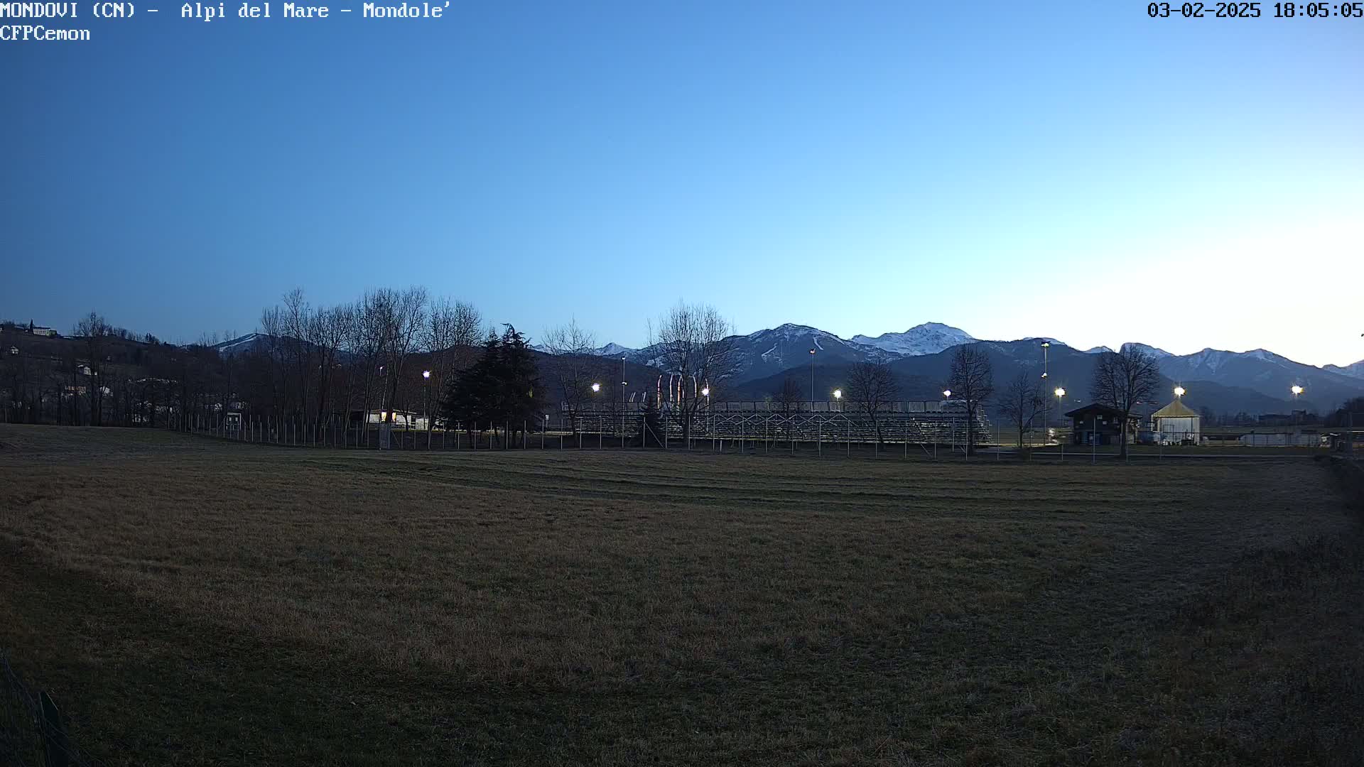 A dark, grassy field sits in front of empty bleachers and a small building, all set against a backdrop of snow-capped mountains under a twilight sky.