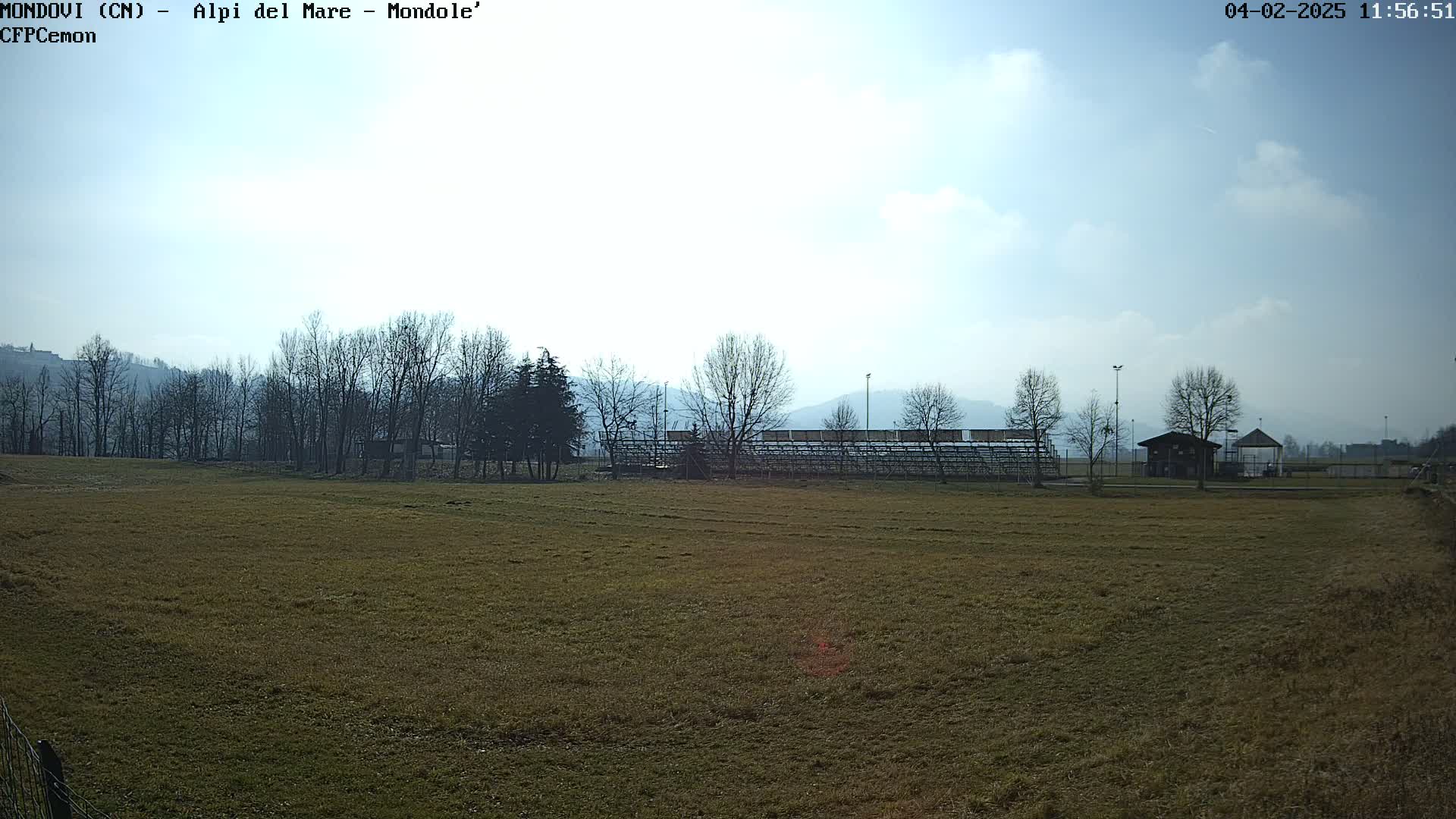 A mostly sunny day reveals a brown grassy field with leafless trees in the background, a small building, and bleachers.