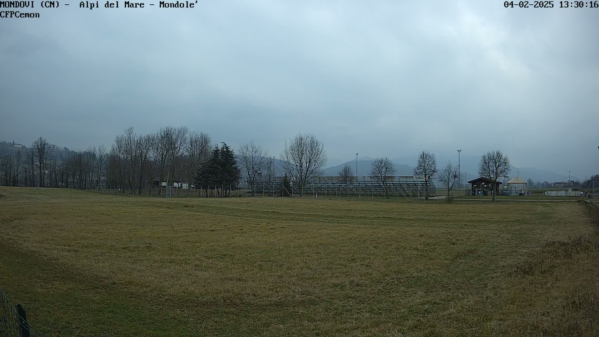 A grassy field stretches towards bleachers and a small building, under an overcast sky, with bare trees and distant mountains visible in the background.