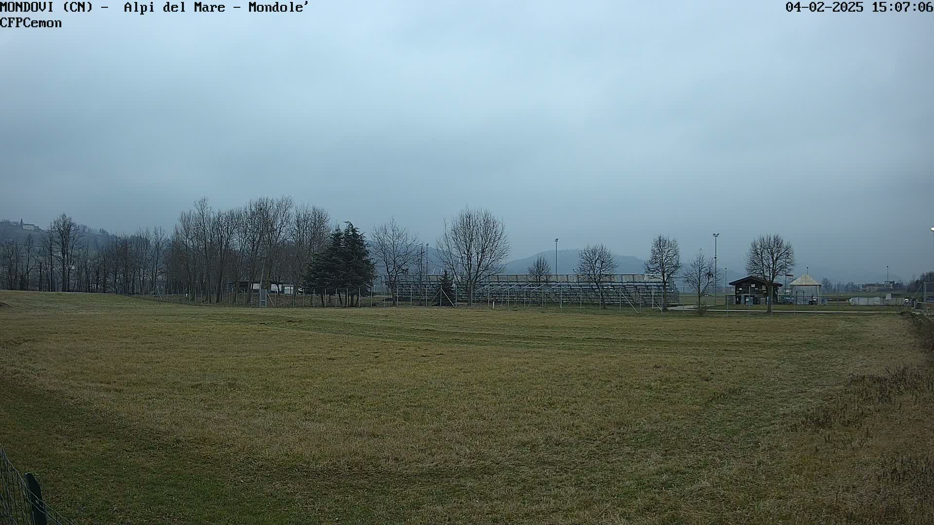 A wide, flat field of brown grass stretches toward bleachers and bare winter trees under an overcast sky.