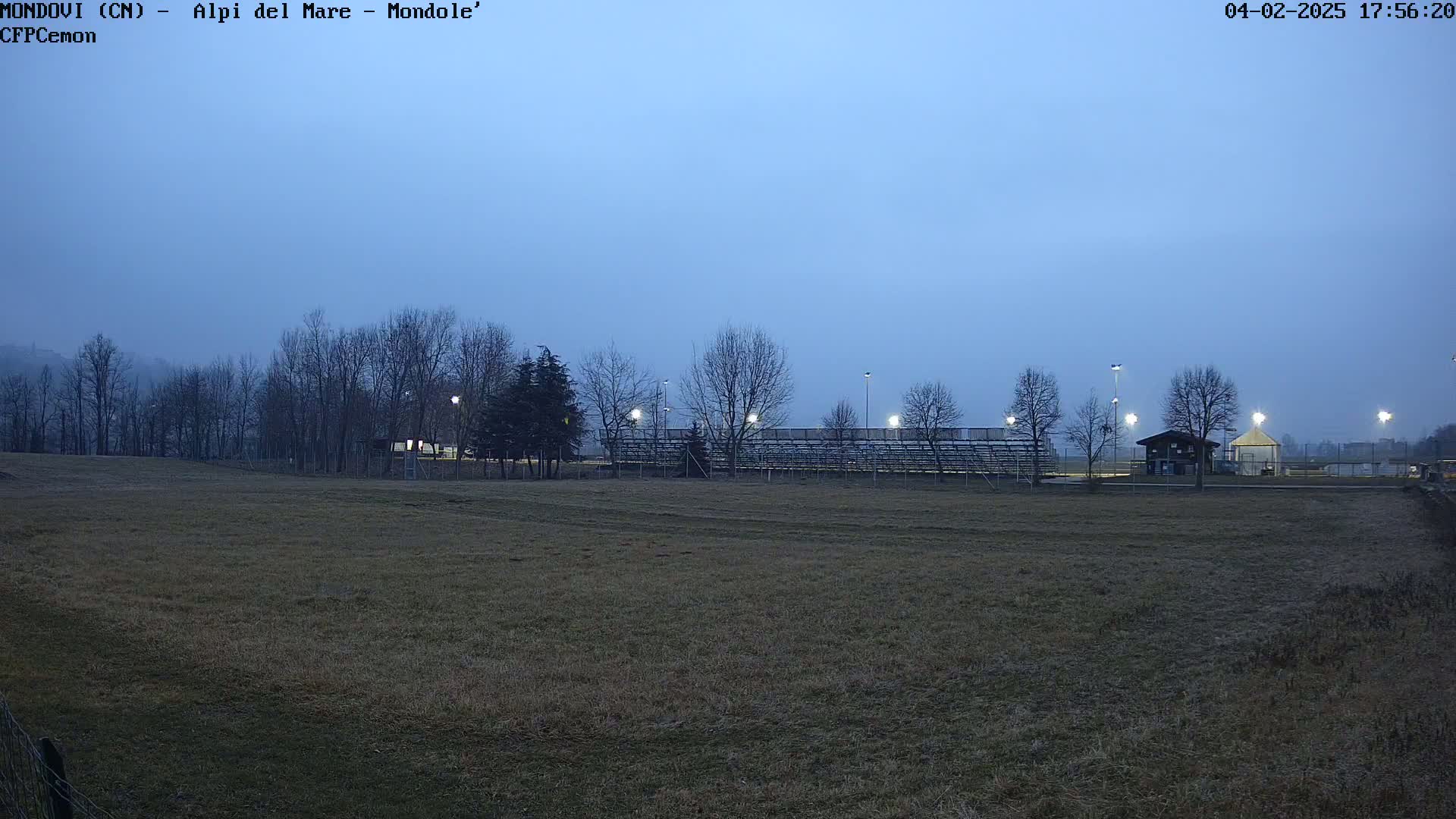 A mostly empty field at dusk is lit by several stadium lights, behind which are bare trees and bleachers.