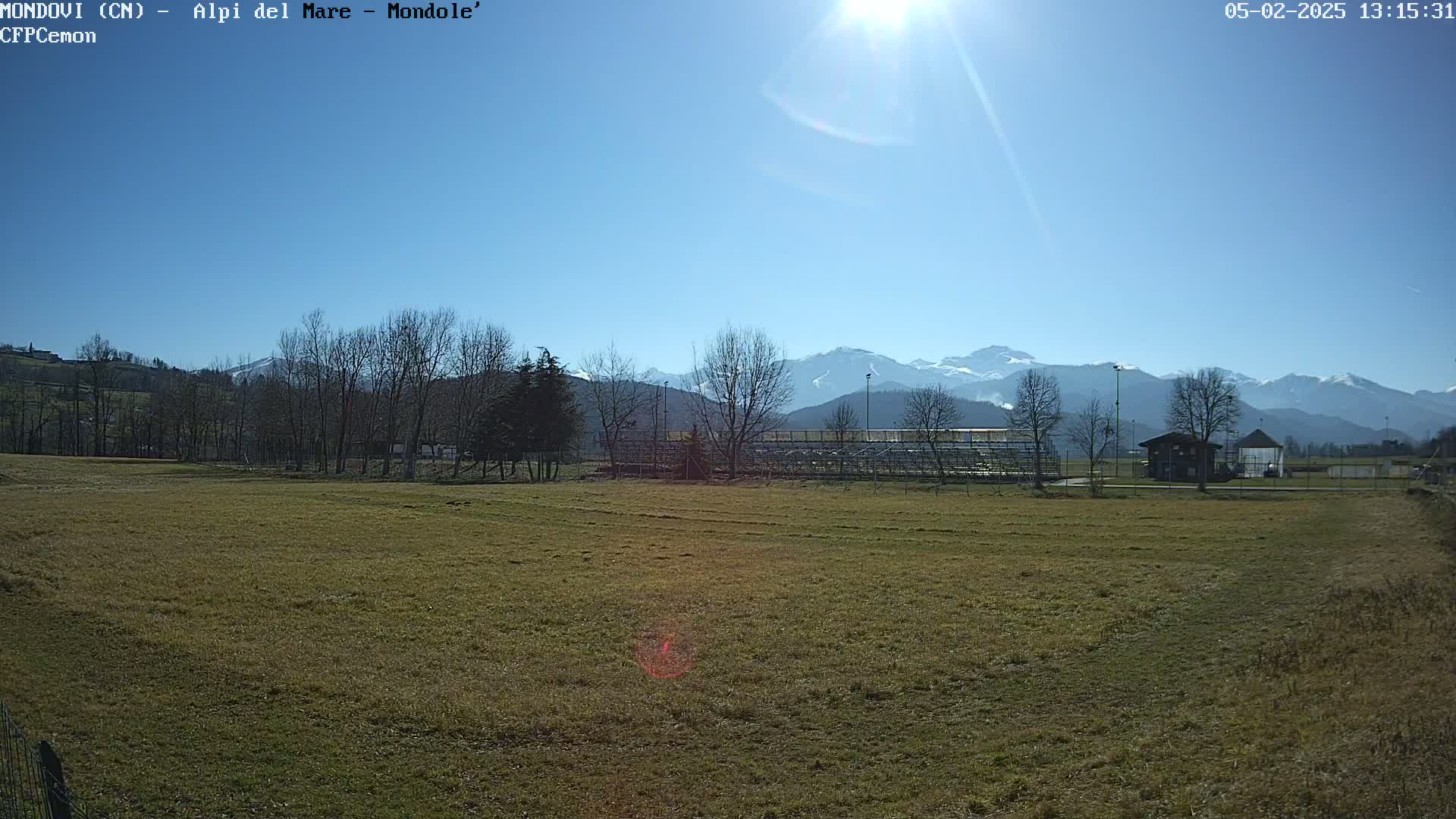 A sunny day reveals a grassy field with bleachers and a small building in the foreground, overlooking a snow-capped mountain range in the background.