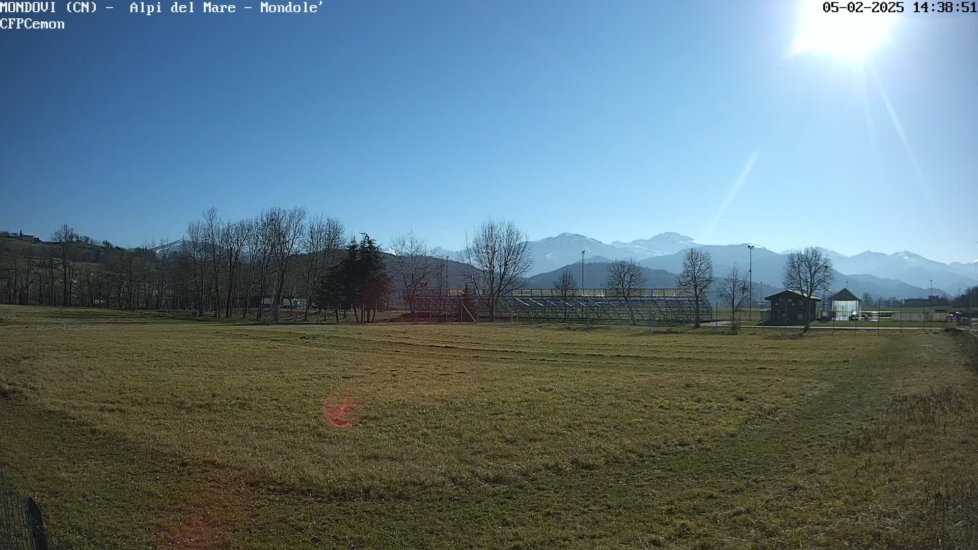 A sunny day reveals a grassy field with bleachers and a small building in the foreground, backed by leafless trees and snow-capped mountains under a clear blue sky.