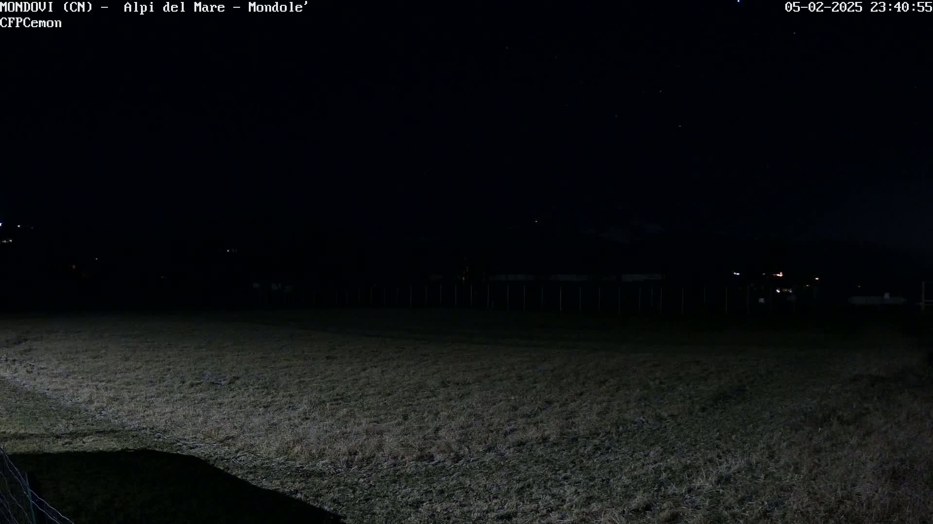 A dark field at night under a clear, starry sky is bordered by a fence and a few distant lights.