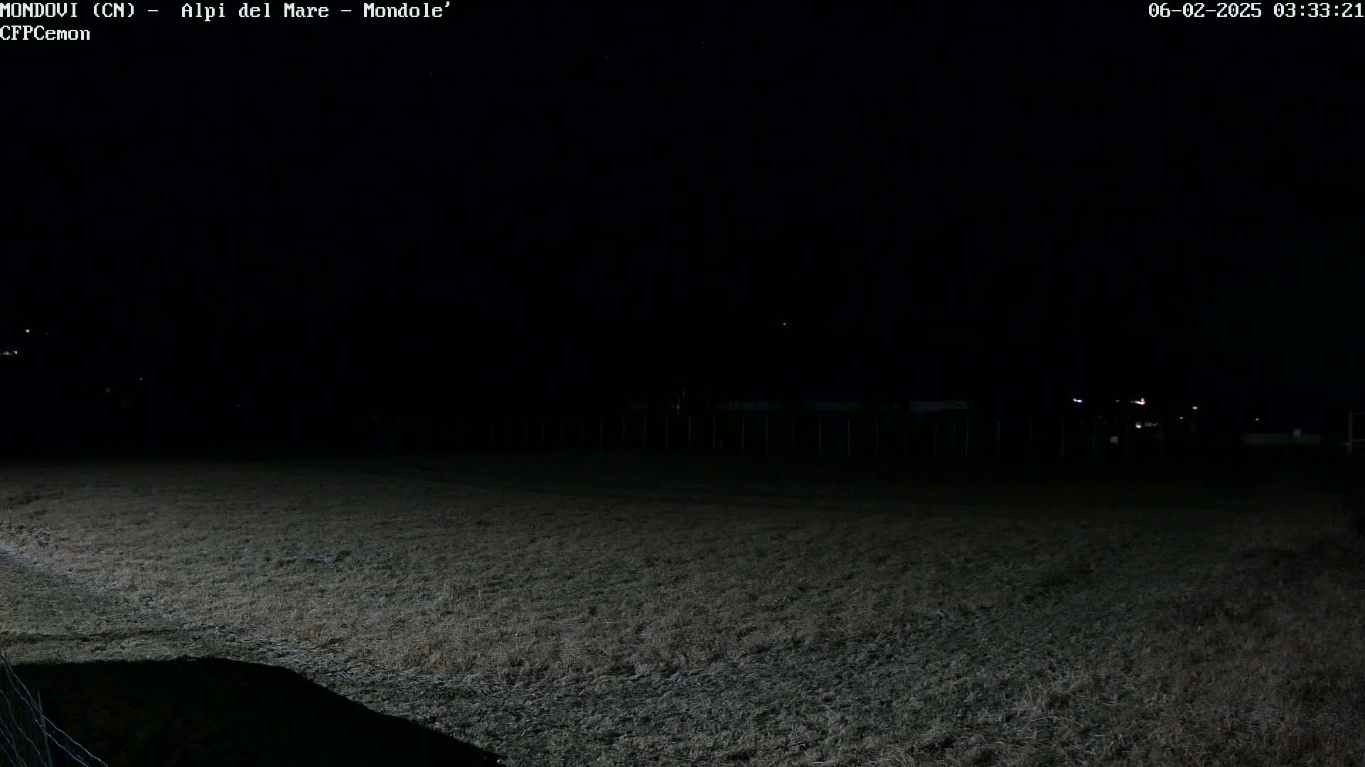 A dark, nighttime view of a grassy field with a fence in the background and a few distant lights visible.