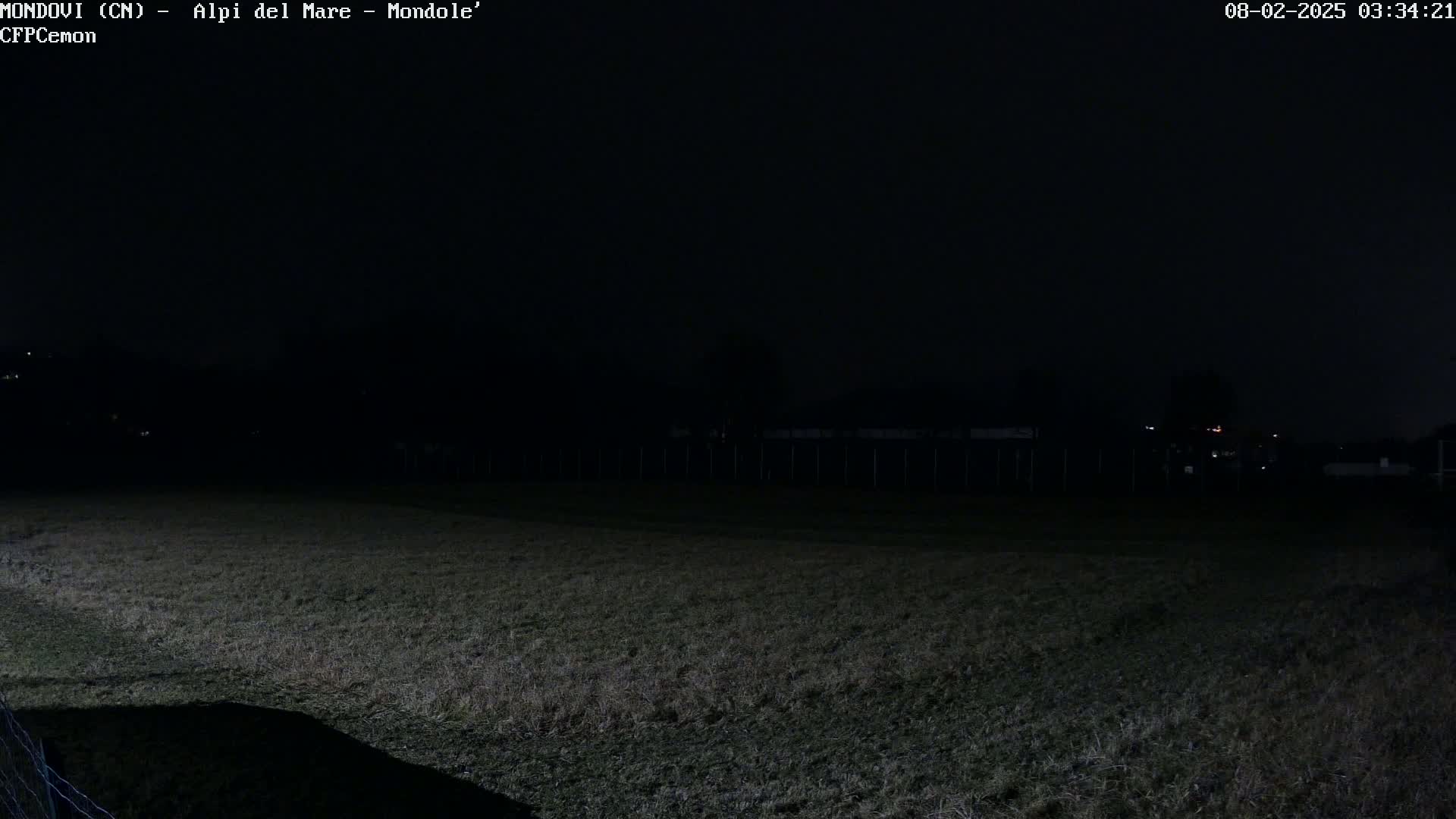 A dark, nighttime view of a grassy field with a fence and dimly lit structures in the background.