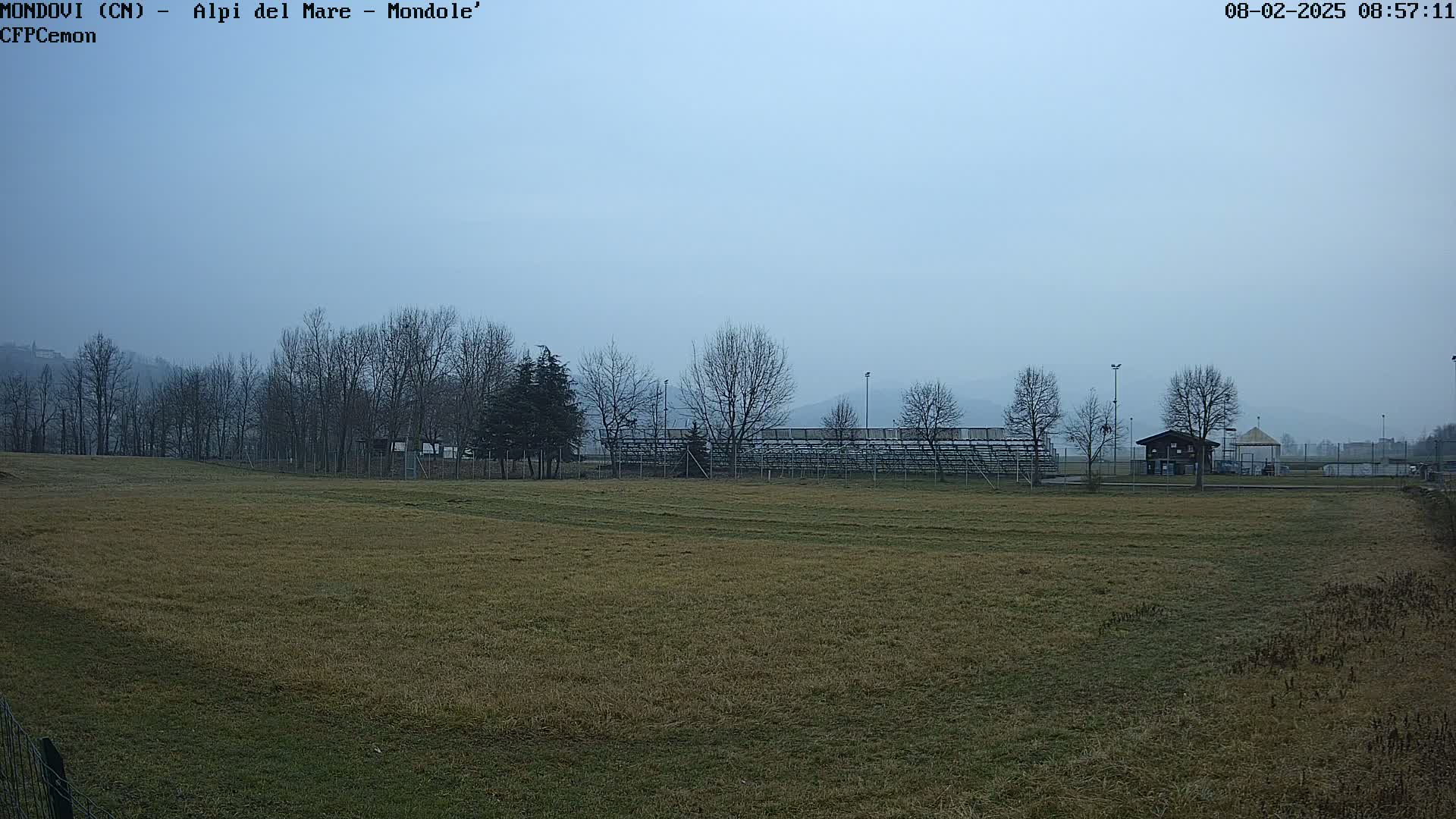 A mostly barren, tan-colored field stretches out before empty bleachers, behind which stand leafless trees under a hazy, overcast sky.