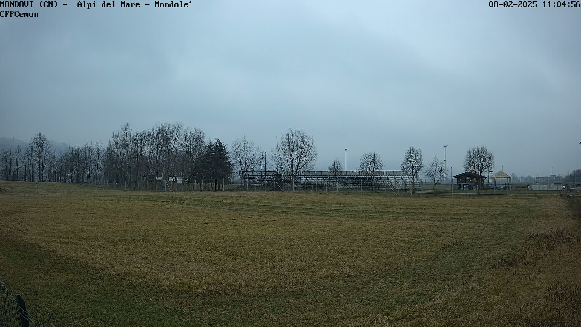 A field with brown grass and bare trees surrounds empty bleachers under an overcast sky.