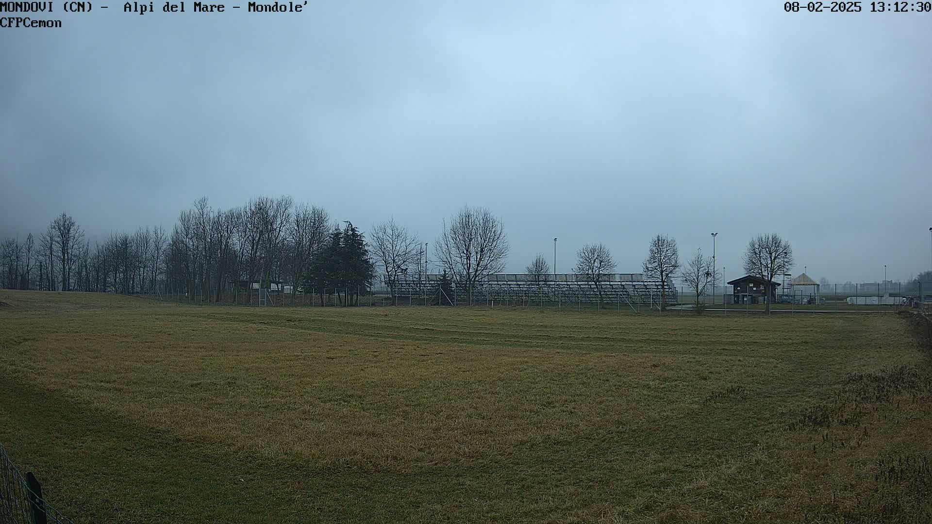 A mostly brown field stretches toward bleachers and bare winter trees under an overcast sky.