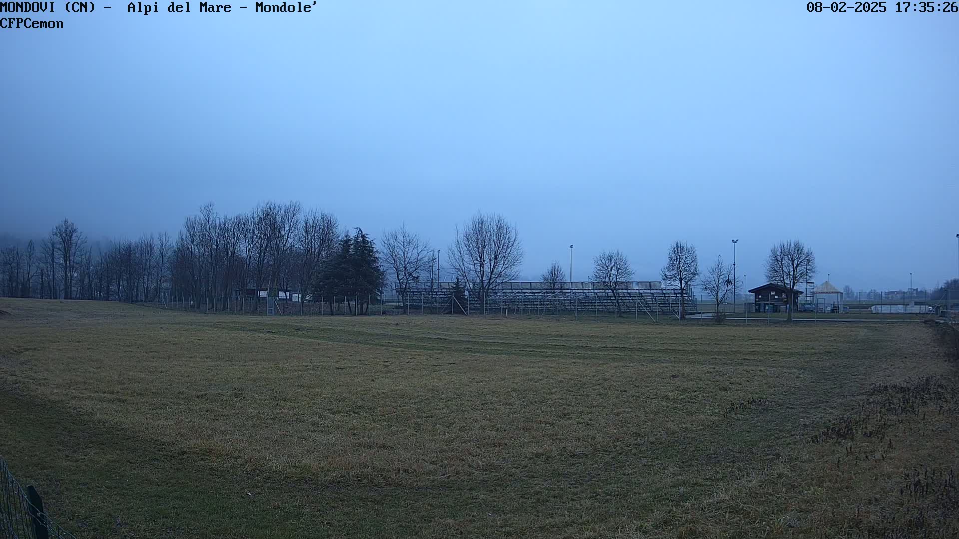 A flat, grassy field stretches toward bleachers and bare winter trees under a hazy, pale blue sky.