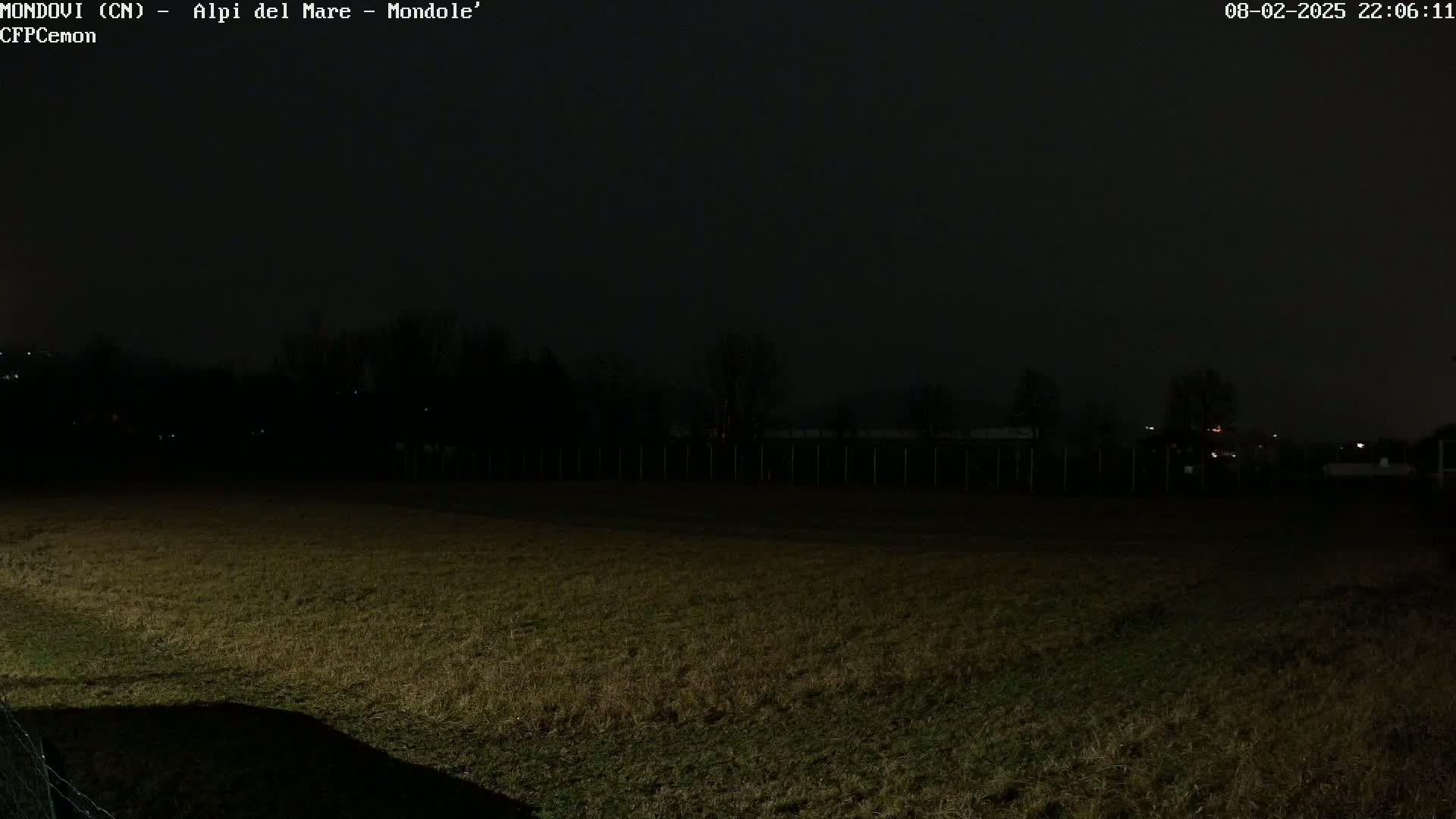 A dark, nighttime view of a grassy field bordered by a fence and trees under a mostly cloudy sky.