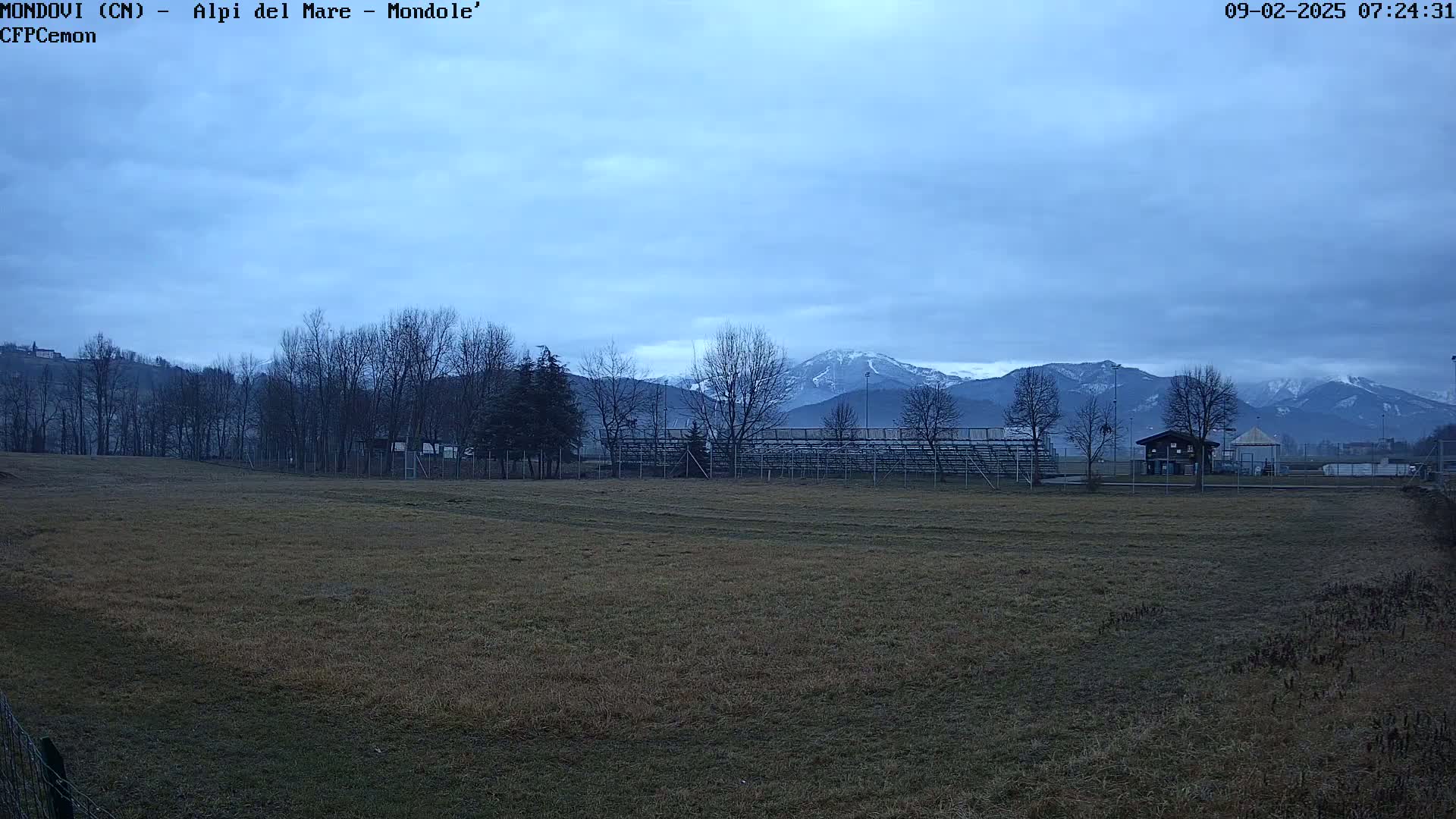 A brown field with bleachers and a small building sits in the foreground, overlooking snow-capped mountains under a cloudy sky.