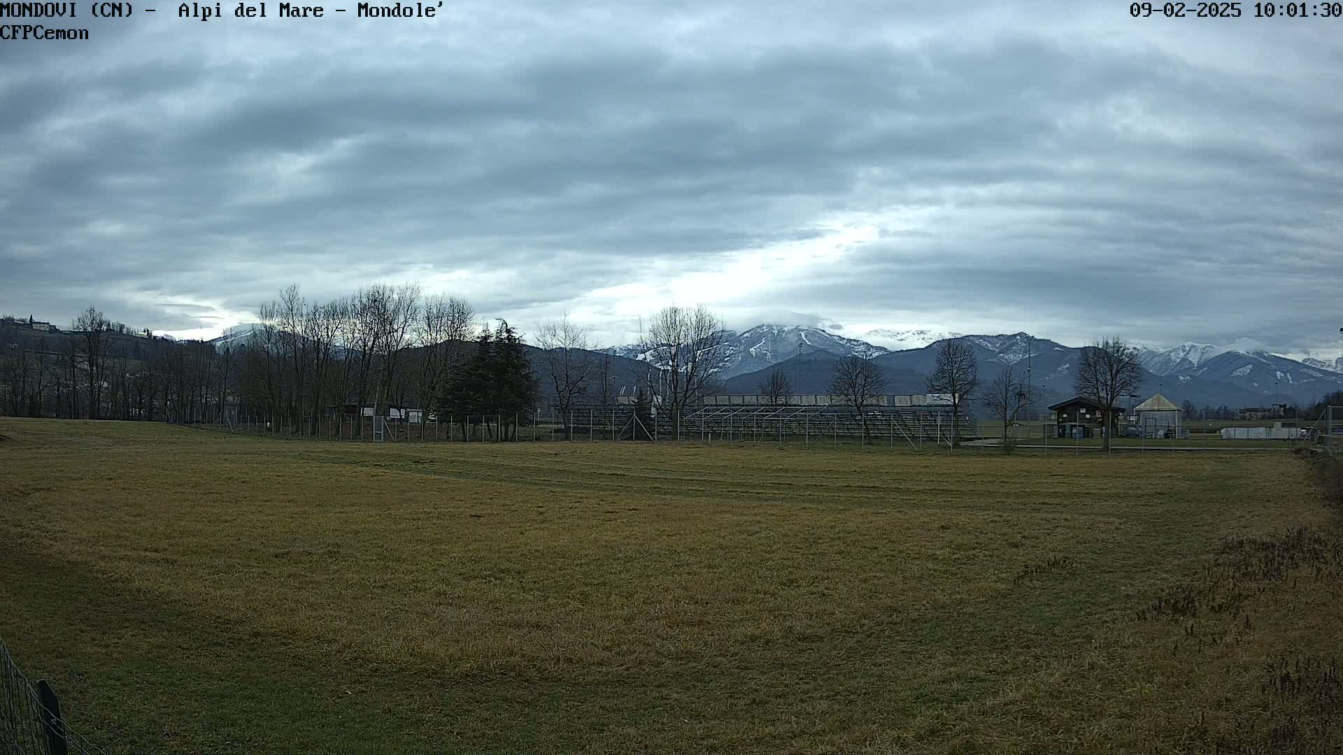 A snow-capped mountain range is visible in the distance beyond a field, bleachers, and bare winter trees under a cloudy sky.