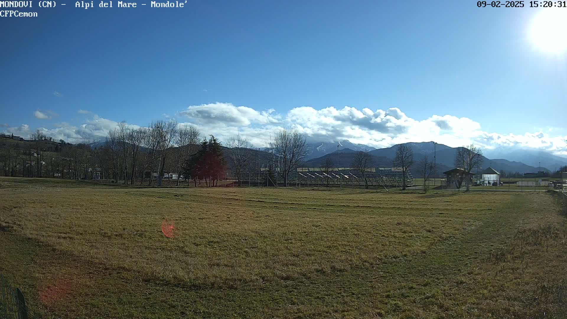 A mostly sunny day reveals a grassy field with a distant empty stadium, bare trees, and snow-capped mountains under a partly cloudy sky.