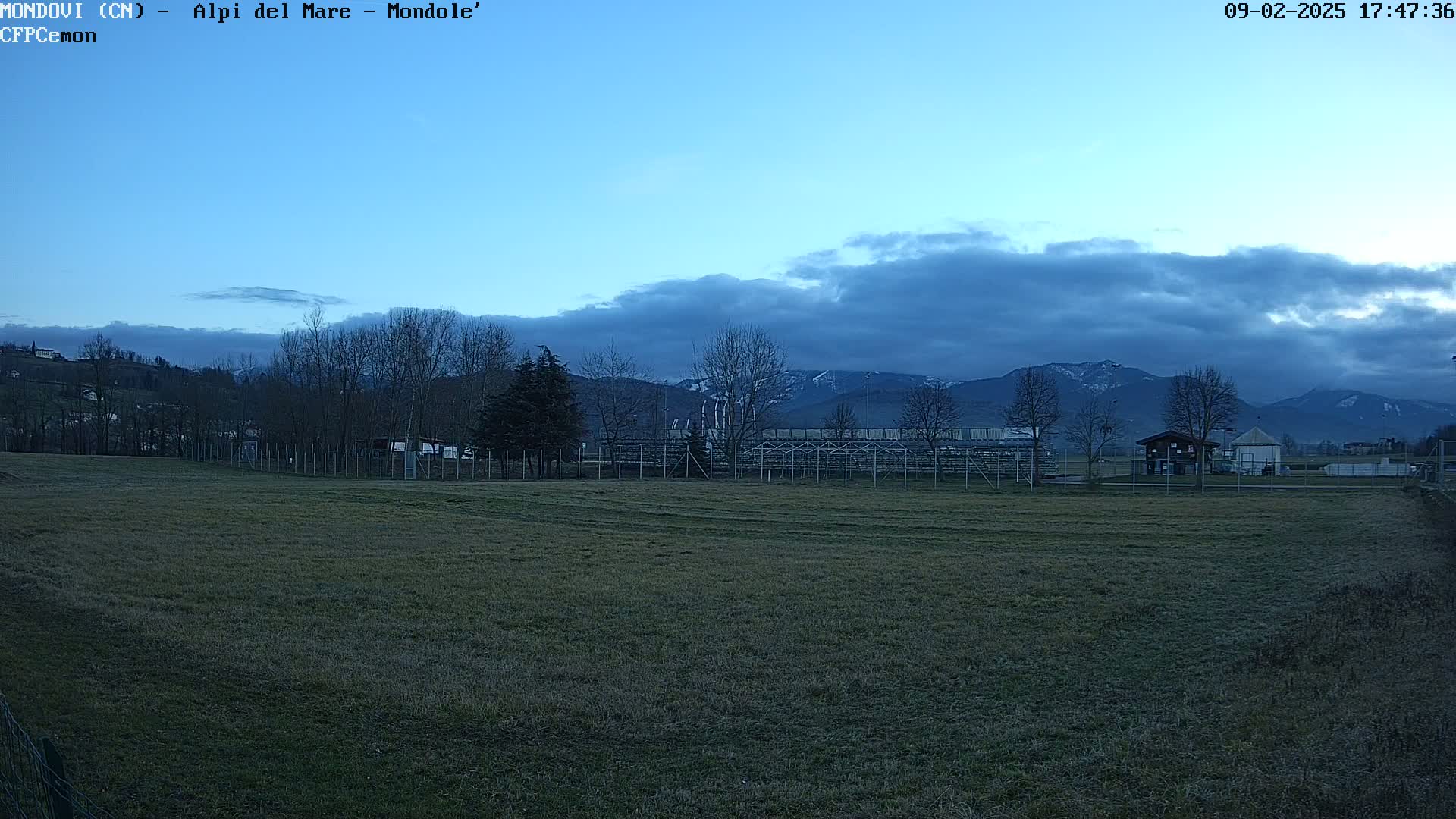 A grassy field with bleachers and a small building sits before a line of bare trees and snow-capped mountains under a mostly cloudy, twilight sky.
