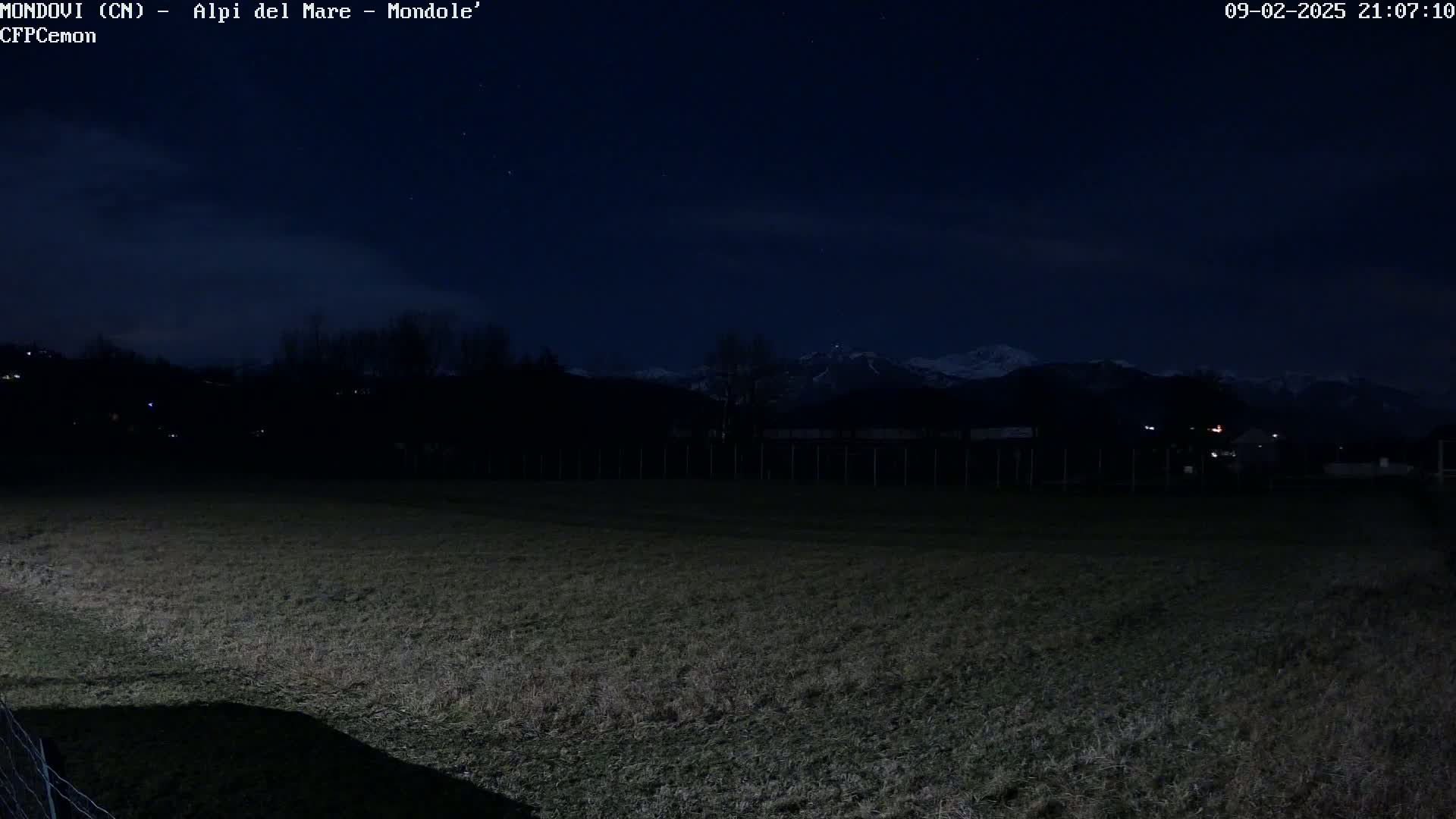 A dark, nighttime field stretches out before snow-capped mountains under a mostly clear, starlit sky.