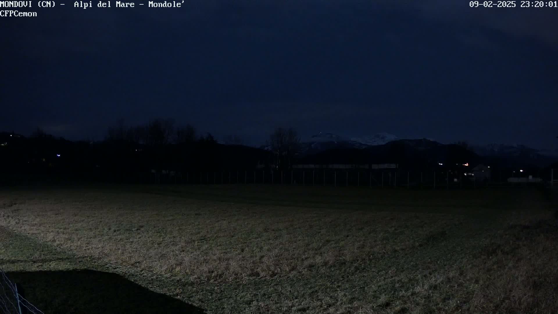 A dark, nighttime view of a grassy field stretches towards a distant, snow-capped mountain range under a clear, dark sky.