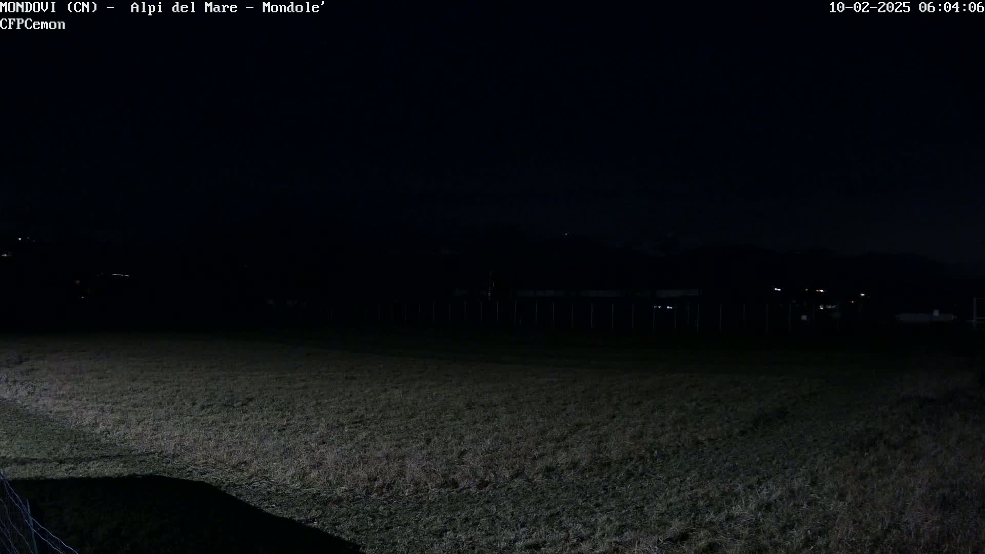 A dark, nighttime view shows a field of grass with a fence and distant lights in the background under a clear, dark sky.