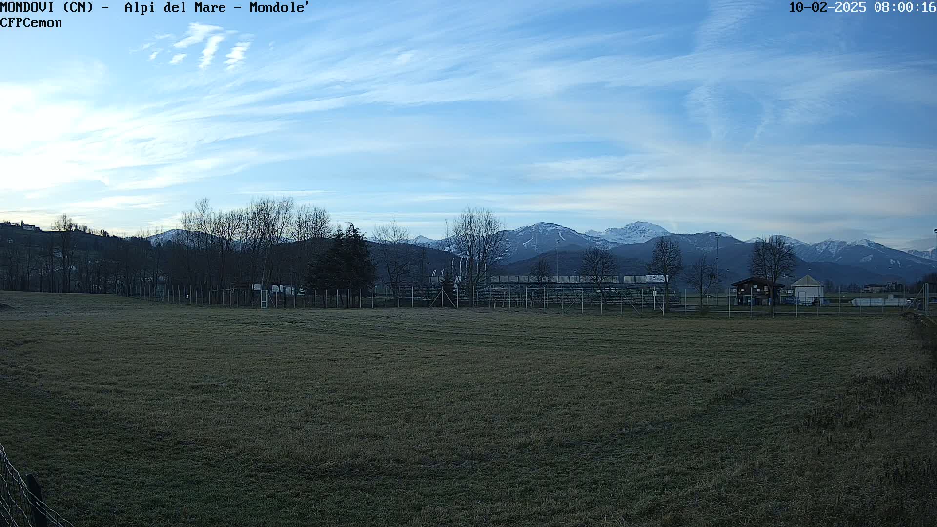 A grassy field with bleachers and a small building sits in the foreground, while snow-capped mountains rise in the background under a mostly clear, light blue sky.