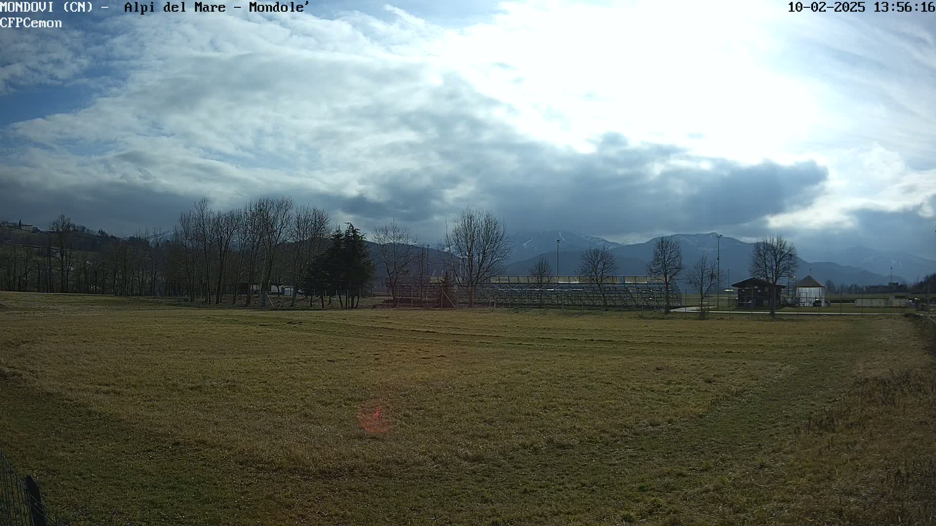 A mostly cloudy sky hangs over a grassy field with bare trees, a small building, and bleachers in the background, with mountains visible in the distance.