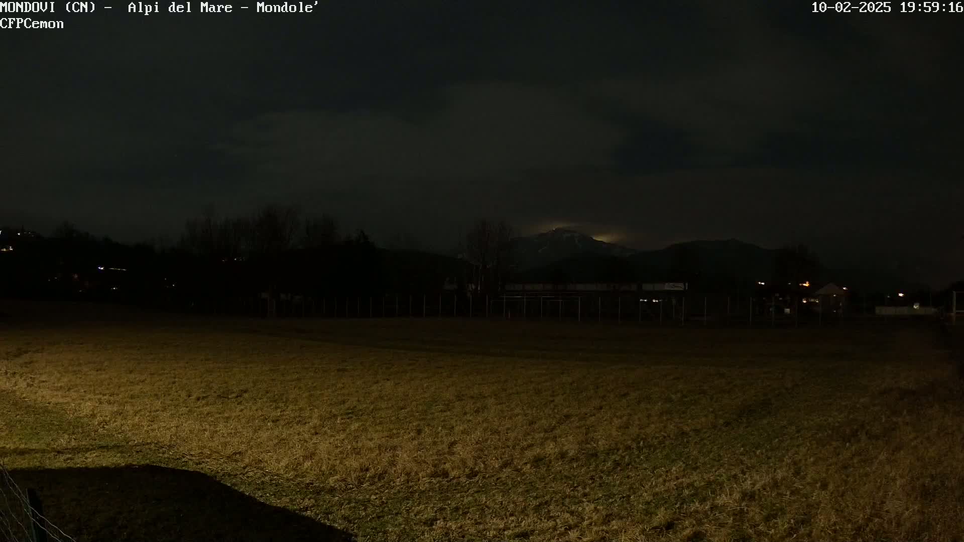 A dark, nighttime view of a grassy field, with a distant mountain range faintly visible under a cloudy sky.