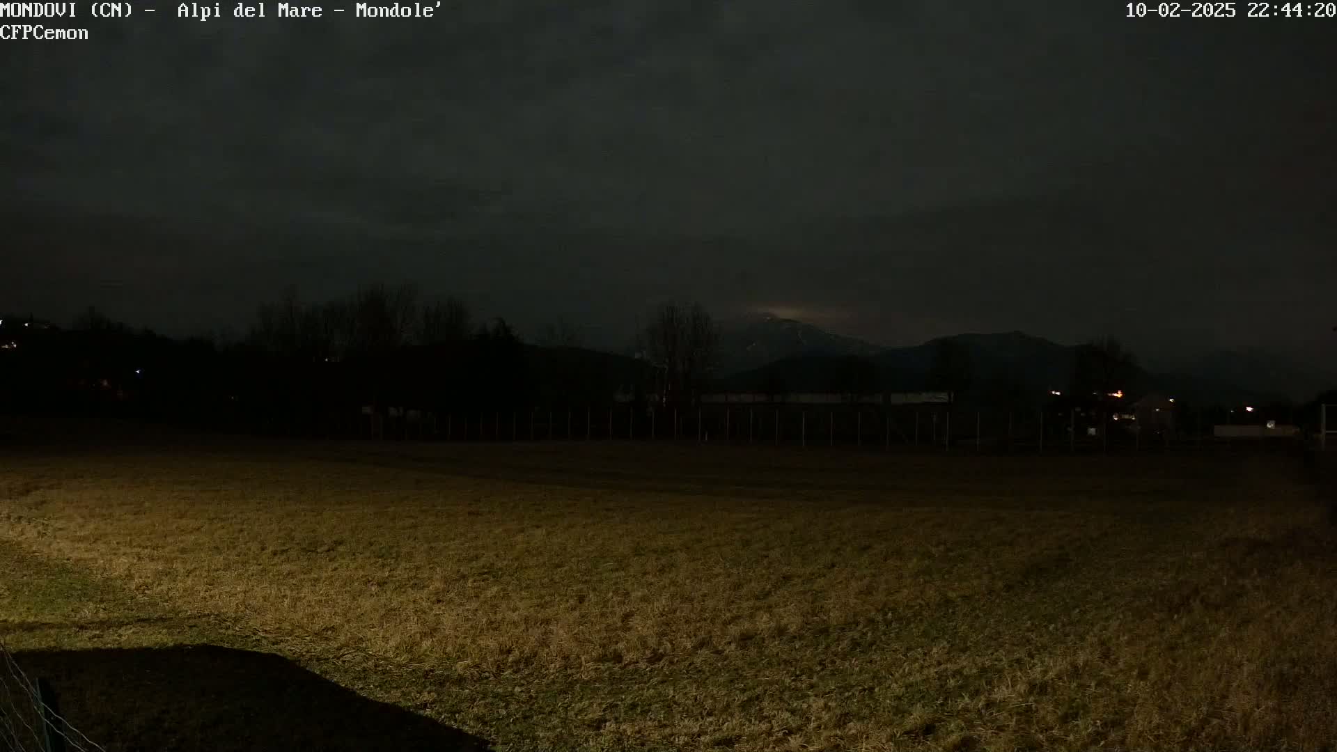 A dark, nighttime field stretches towards a dimly lit mountain range under a mostly cloudy sky.