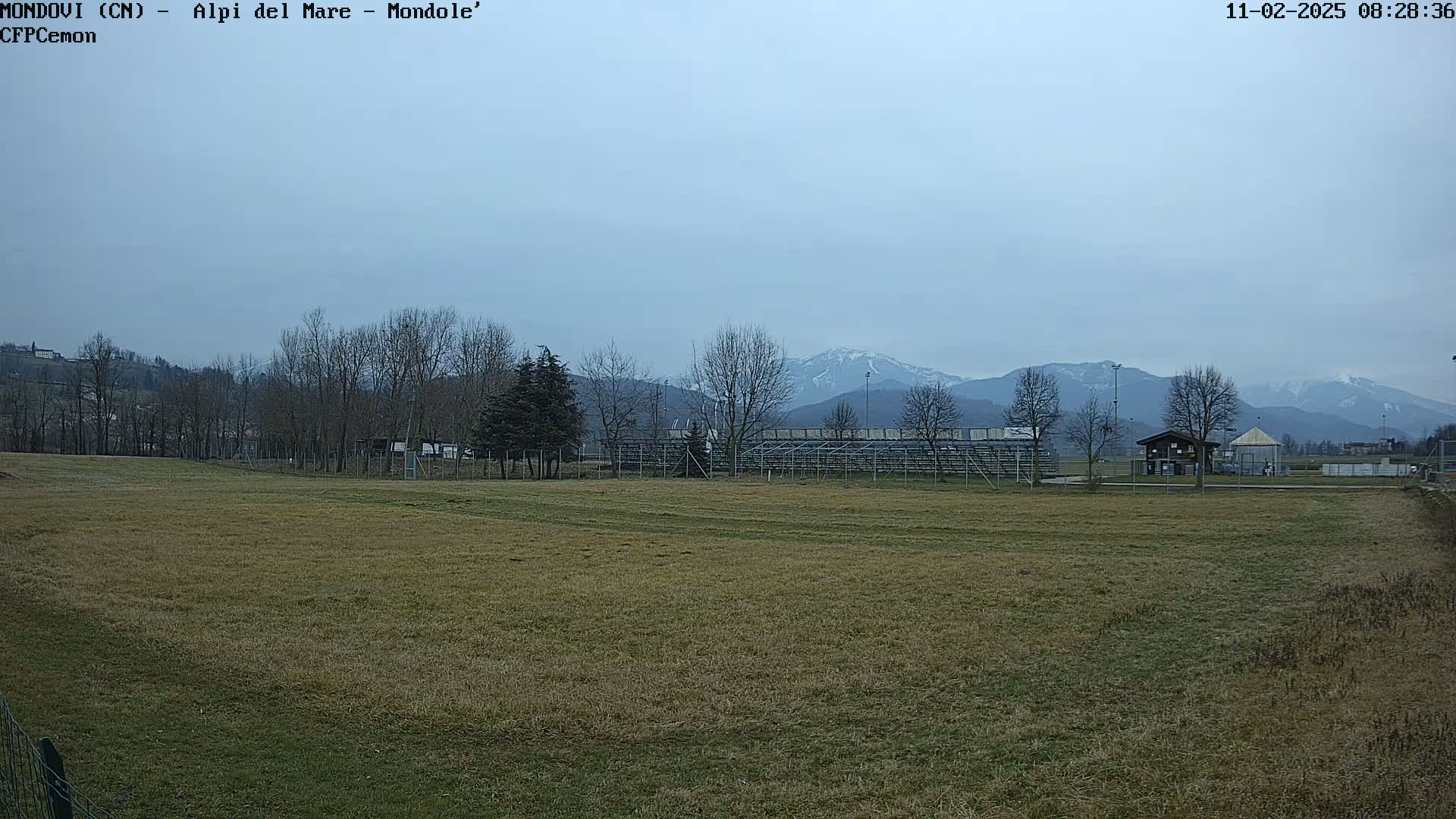 A mostly brown field stretches toward bleachers and a small building, with bare trees and snow-capped mountains in the background under a cloudy sky.