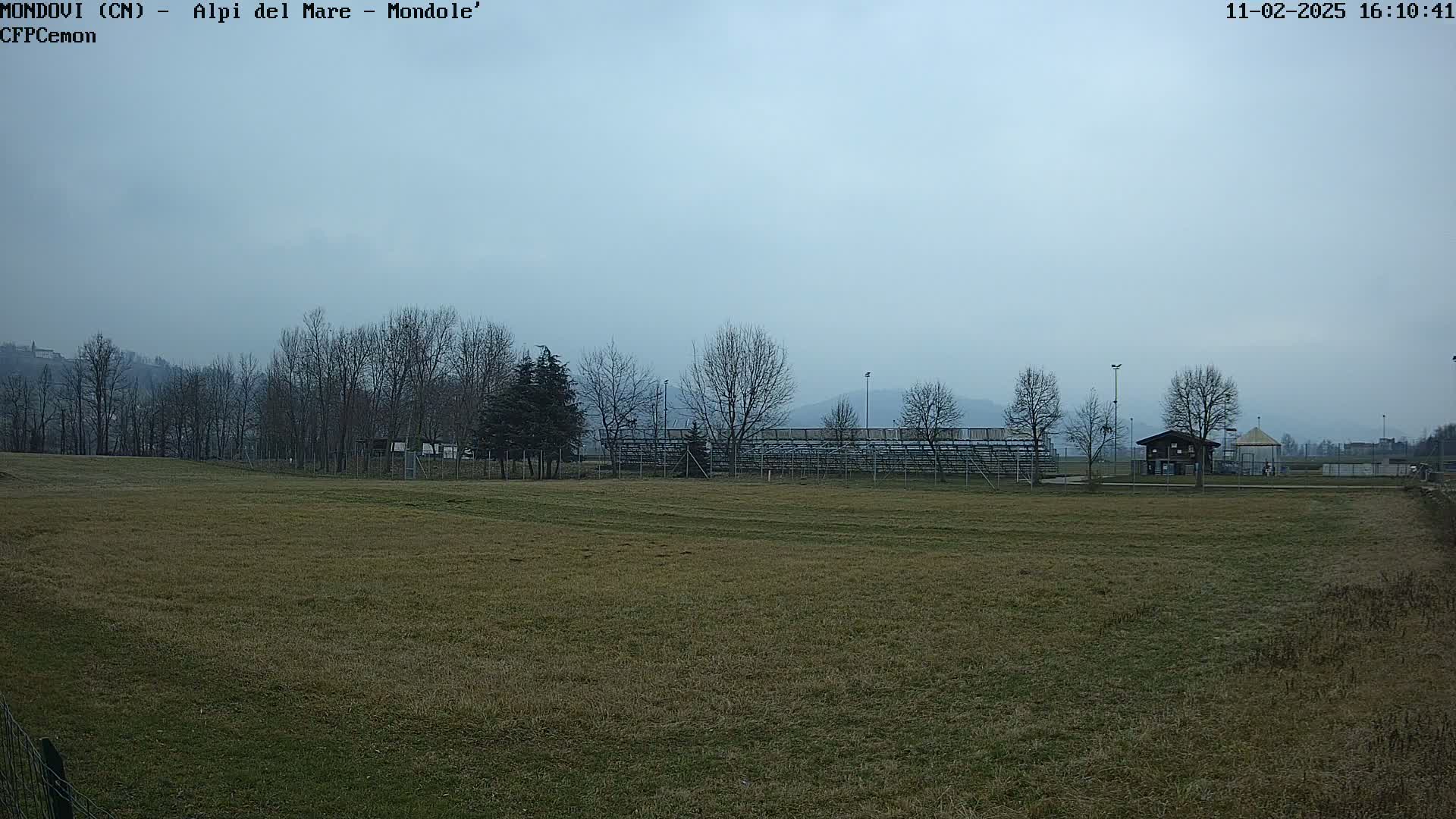 A grassy field with bleachers and a few buildings in the background under a hazy, overcast sky.