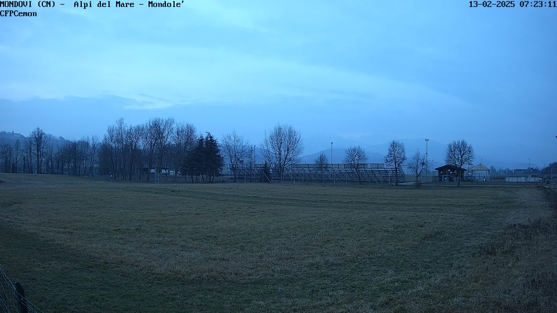 A mostly barren field with bleachers and a small building is visible under a pale blue, overcast sky, with bare trees and distant mountains in the background.