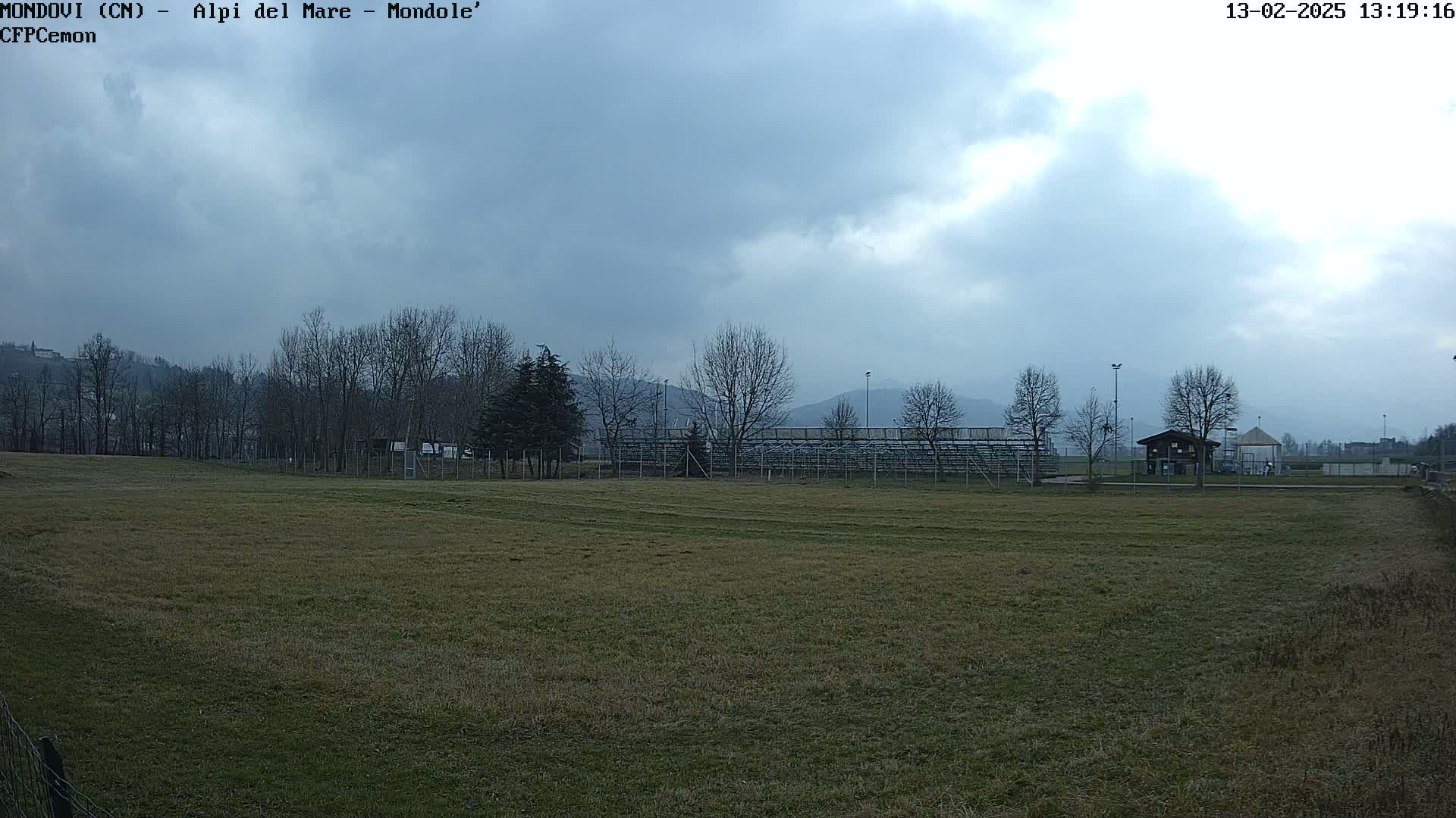 A mostly cloudy day reveals a grassy field with bleachers and a small building in the background, overlooked by bare trees and distant mountains.