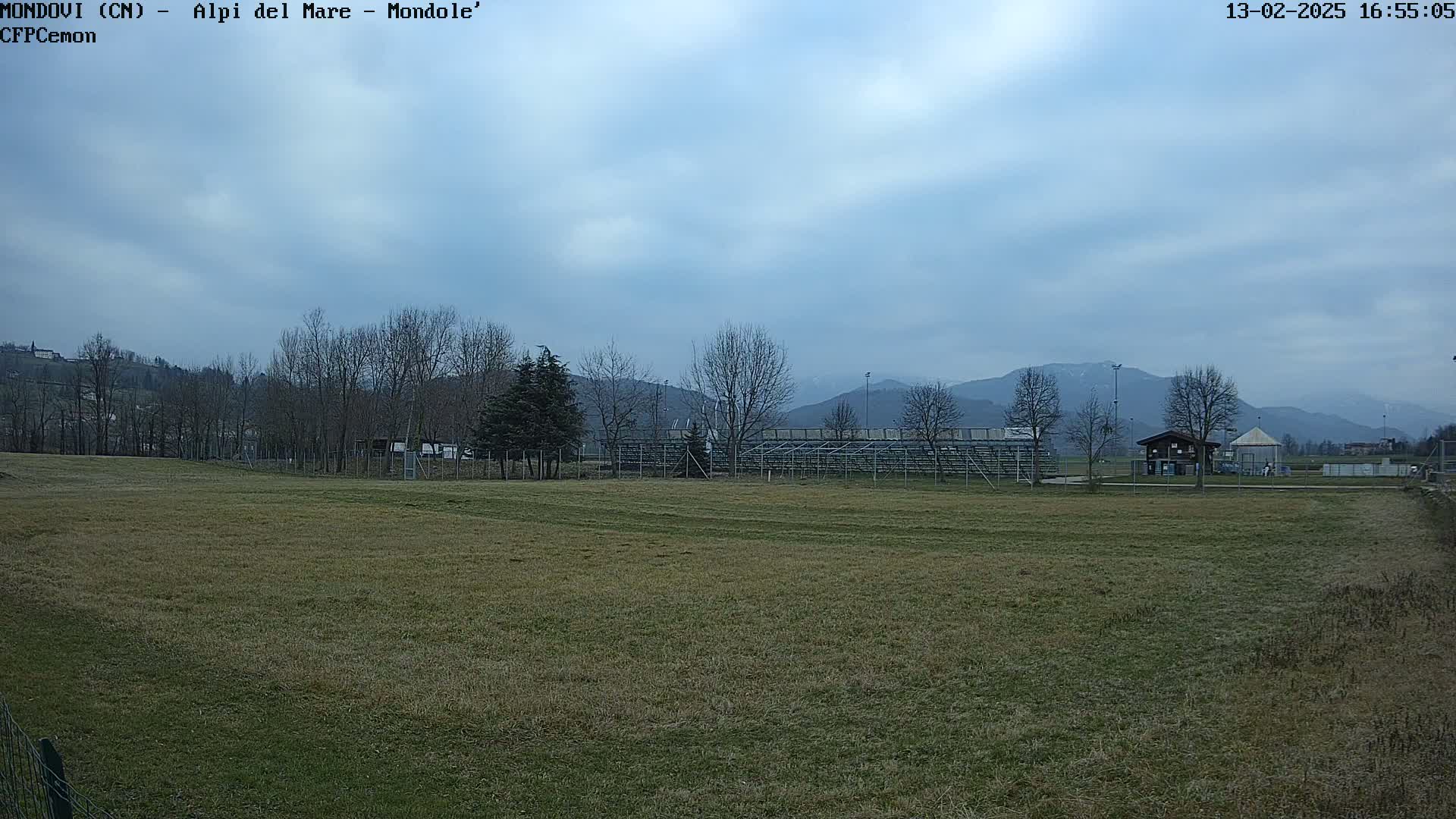 A mostly cloudy day reveals a grassy field with bleachers and a small building in the background, mountains visible in the distance.