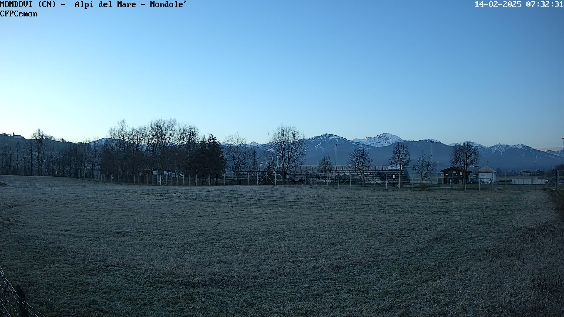A frost-covered field stretches towards a line of leafless trees and snow-capped mountains under a clear, twilight sky.